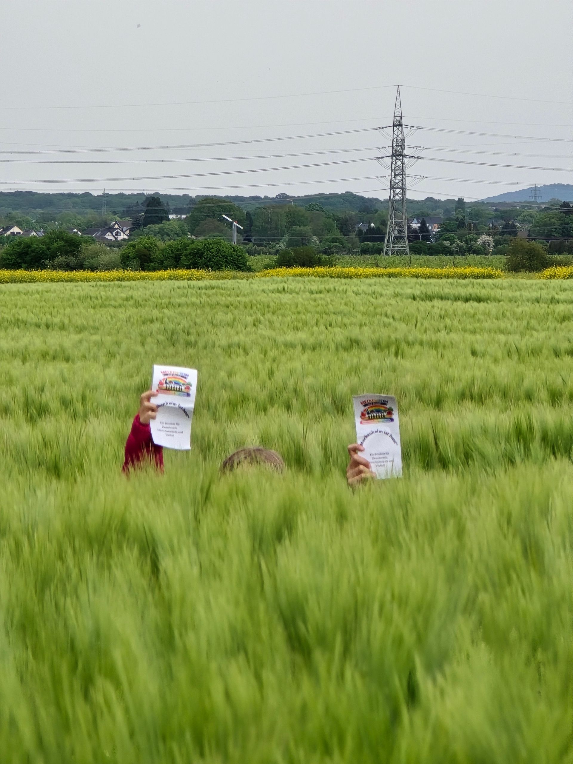 Zwei Personen halten Flyer auf einen Gerstenfeld; im Hintergrund sind Stromleitungen und Hügel zu sehen.
