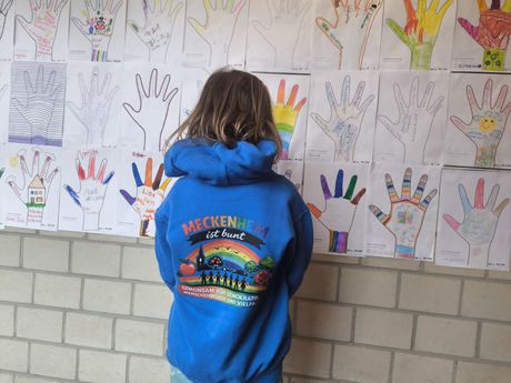 Ein Kind in einem blauen Kapuzenpulli steht vor einer Wand, die mit bunten, handgezeichneten Handabdrücken bedeckt ist.