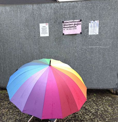 Ein Regenbogen-Regenschirm vor einer grauen Wand mit einigen Aushängen.