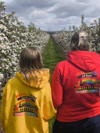 Zwei Personen in gelben und roten Kapuzenpullovern gehen unter bewölktem Himmel einen blumengesäumten Weg entlang.