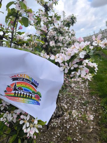 Ein weißer Rucksack mit farbenfrohem Logo, gehalten zwischen rosa Frühlingsblüten auf einem von Bäumen gesäumten Weg
