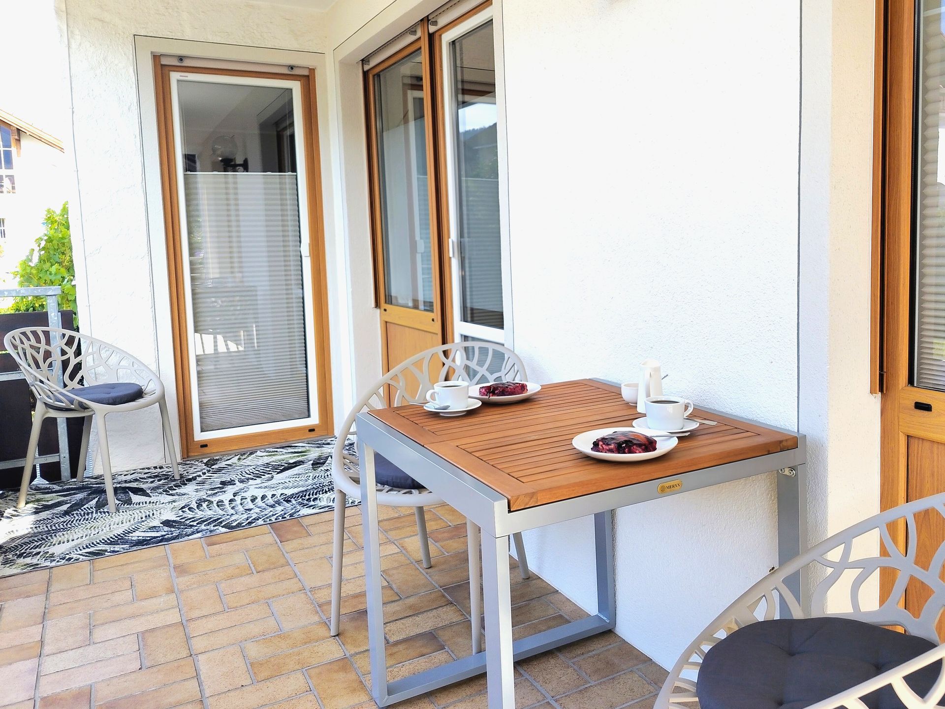 Terrasse mit gedecktem Holztisch für Tee neben einer Glastür und Stuhl auf Fliesenboden.