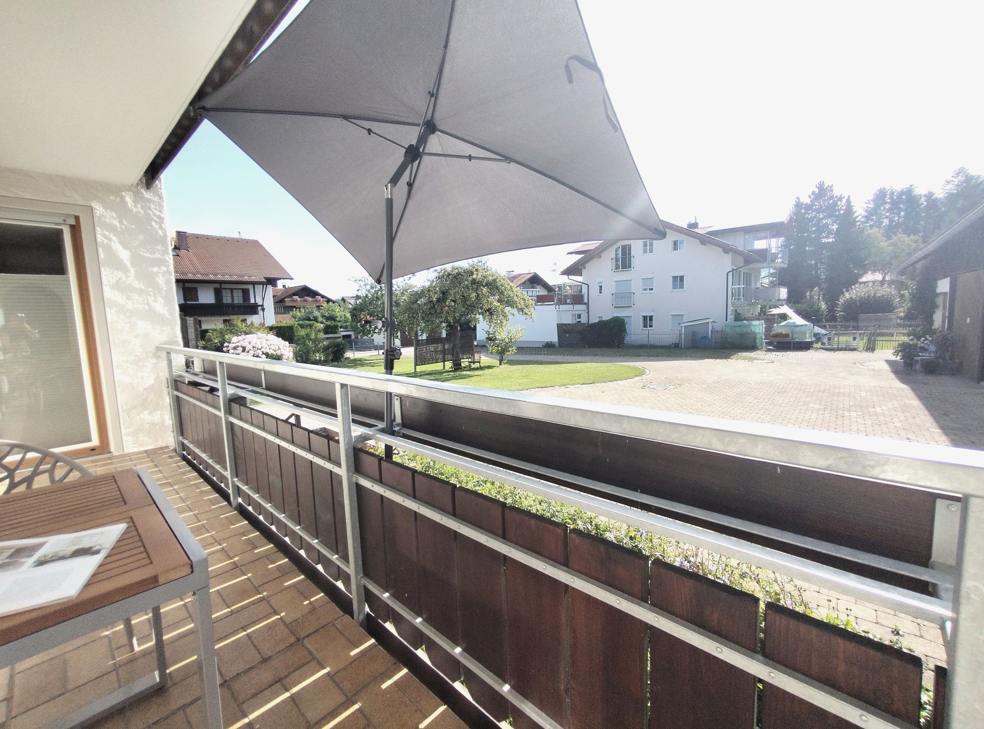 Balkon mit Fliesenboden, Geländer und großem Sonnenschirm mit Blick auf weiße Häuser und eine sonnige Straße