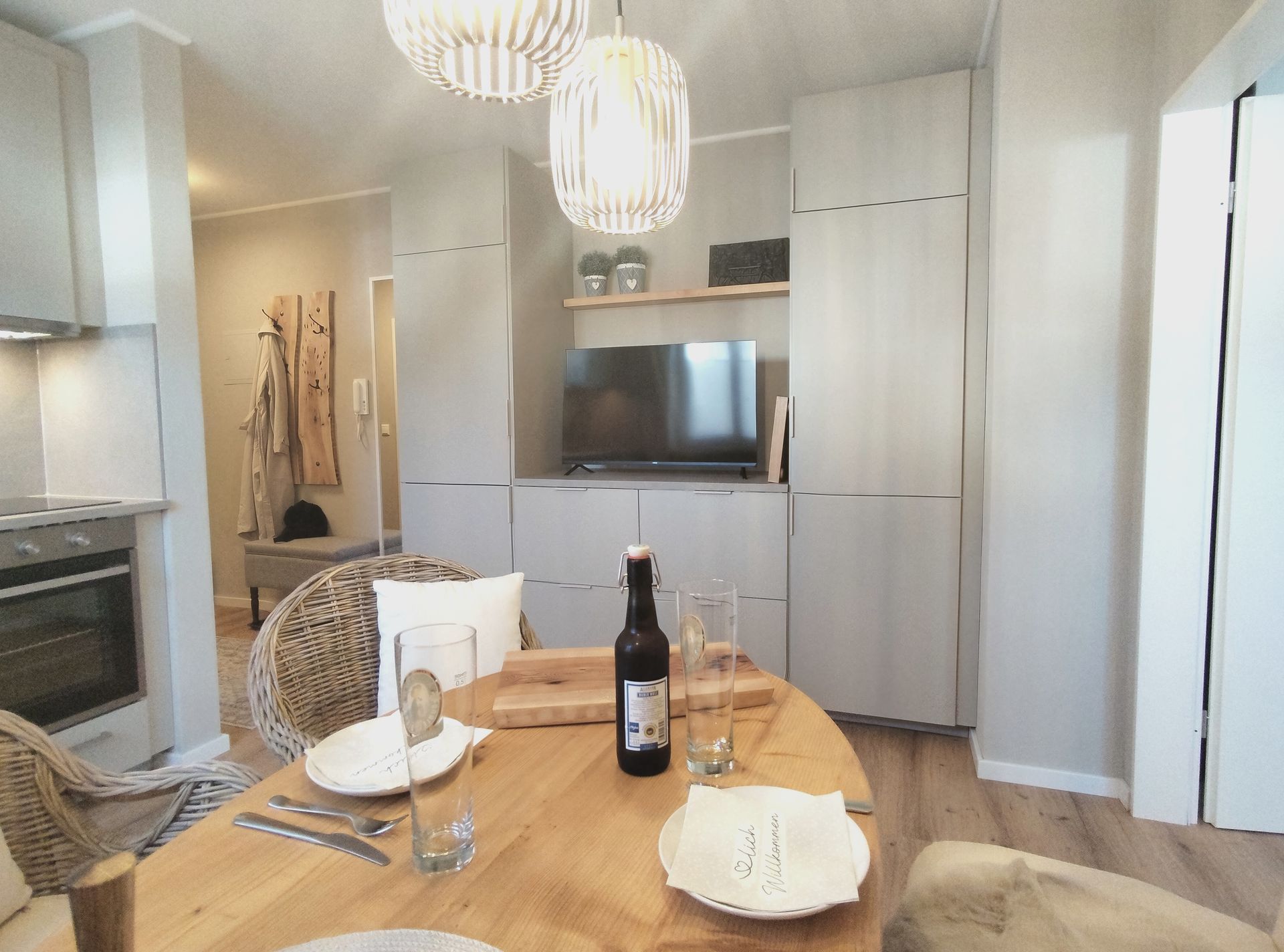 Esstisch in einem hellen, modernen Wohnzimmer mit weißen Schränken, Fernseher, Kronleuchter und Weinflaschen-Mittelstück