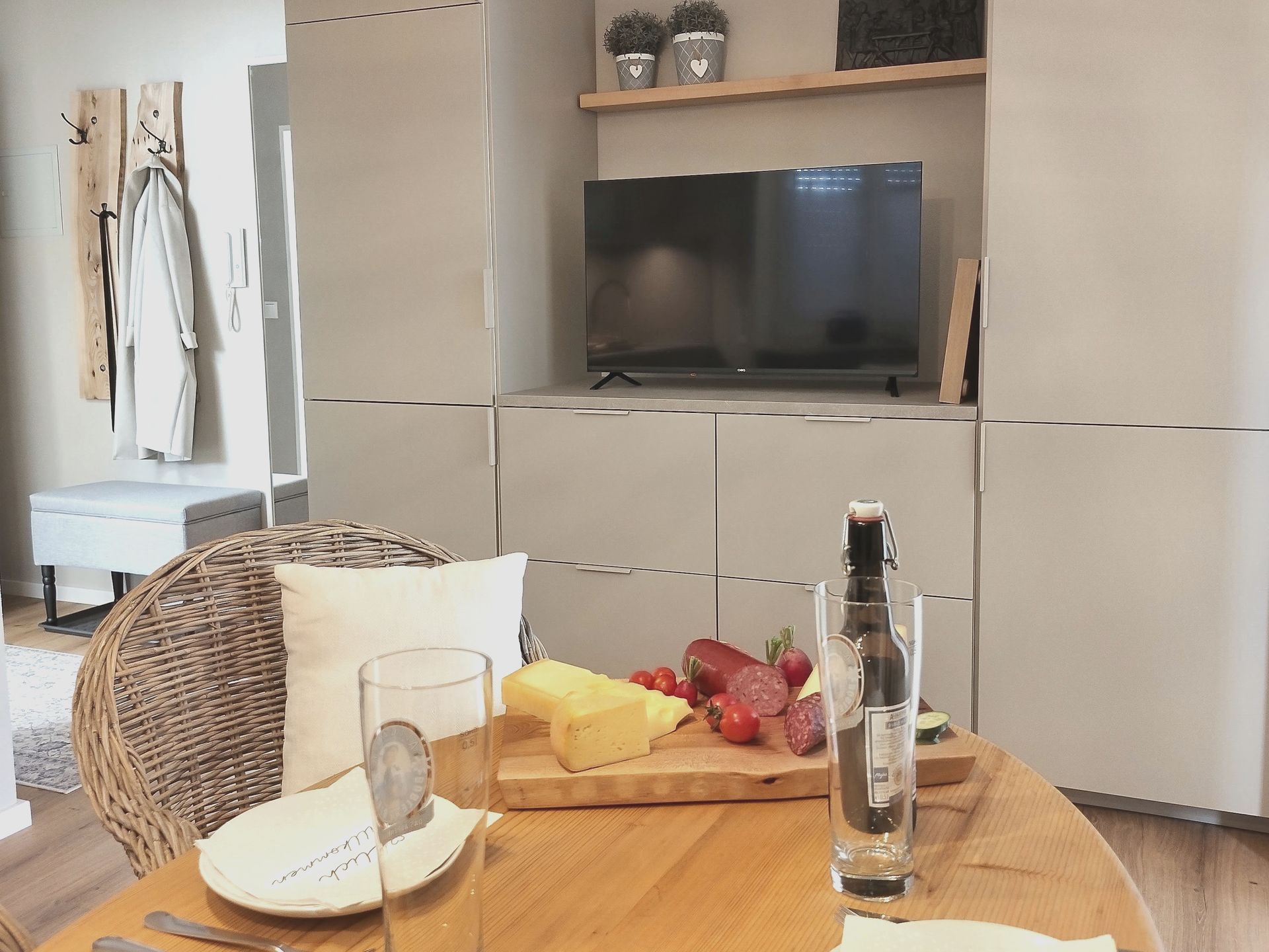 Gemütliches Wohnzimmer mit Fernseher, einem Holztablett mit Obst und Käse auf einem Tisch und einem Korbsessel im Vordergrund