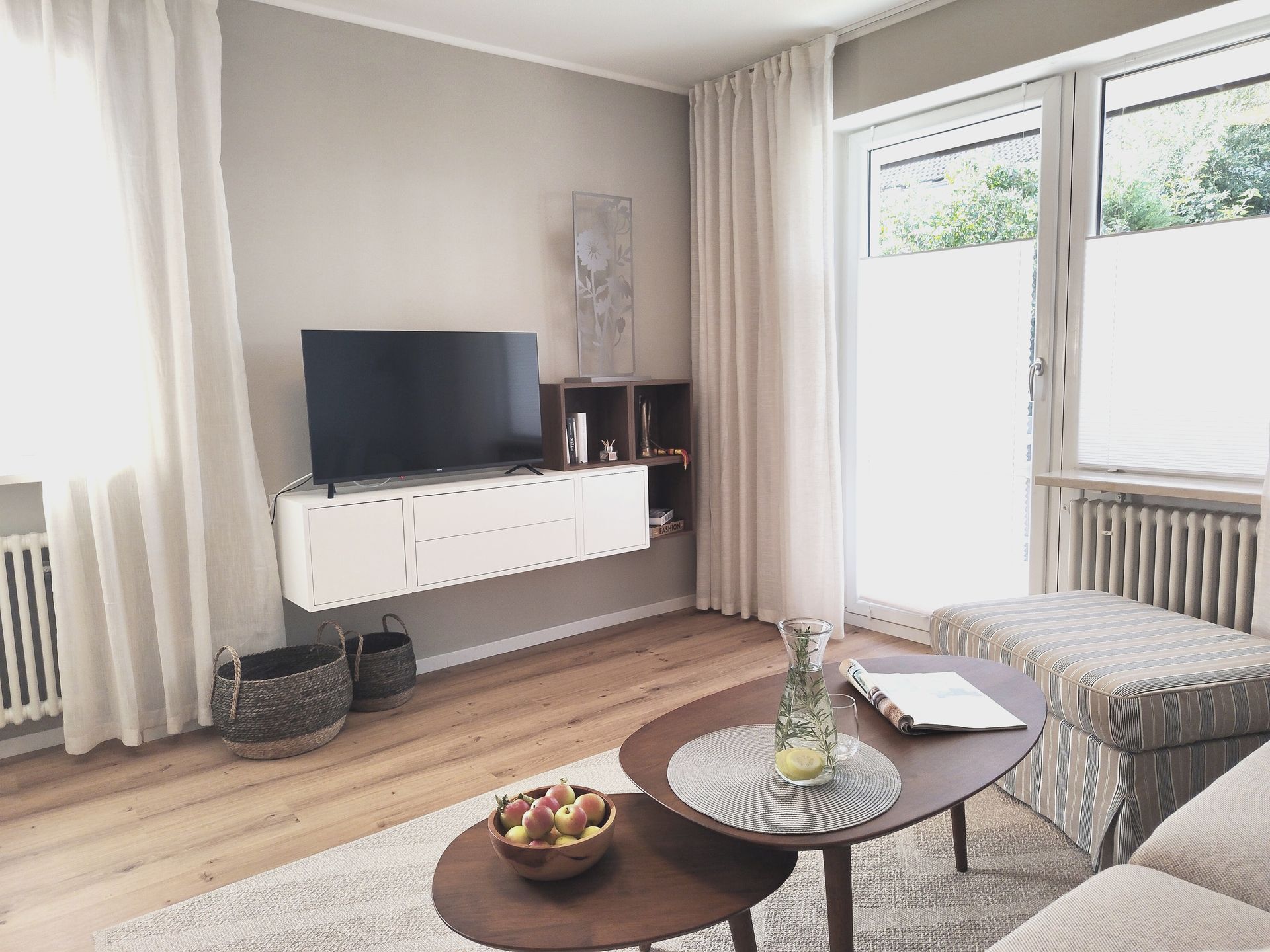 Helles Wohnzimmer mit Fernseher, weißem Schrank, großen Fenstern und rundem Couchtisch mit Obst und Zeitschriften.