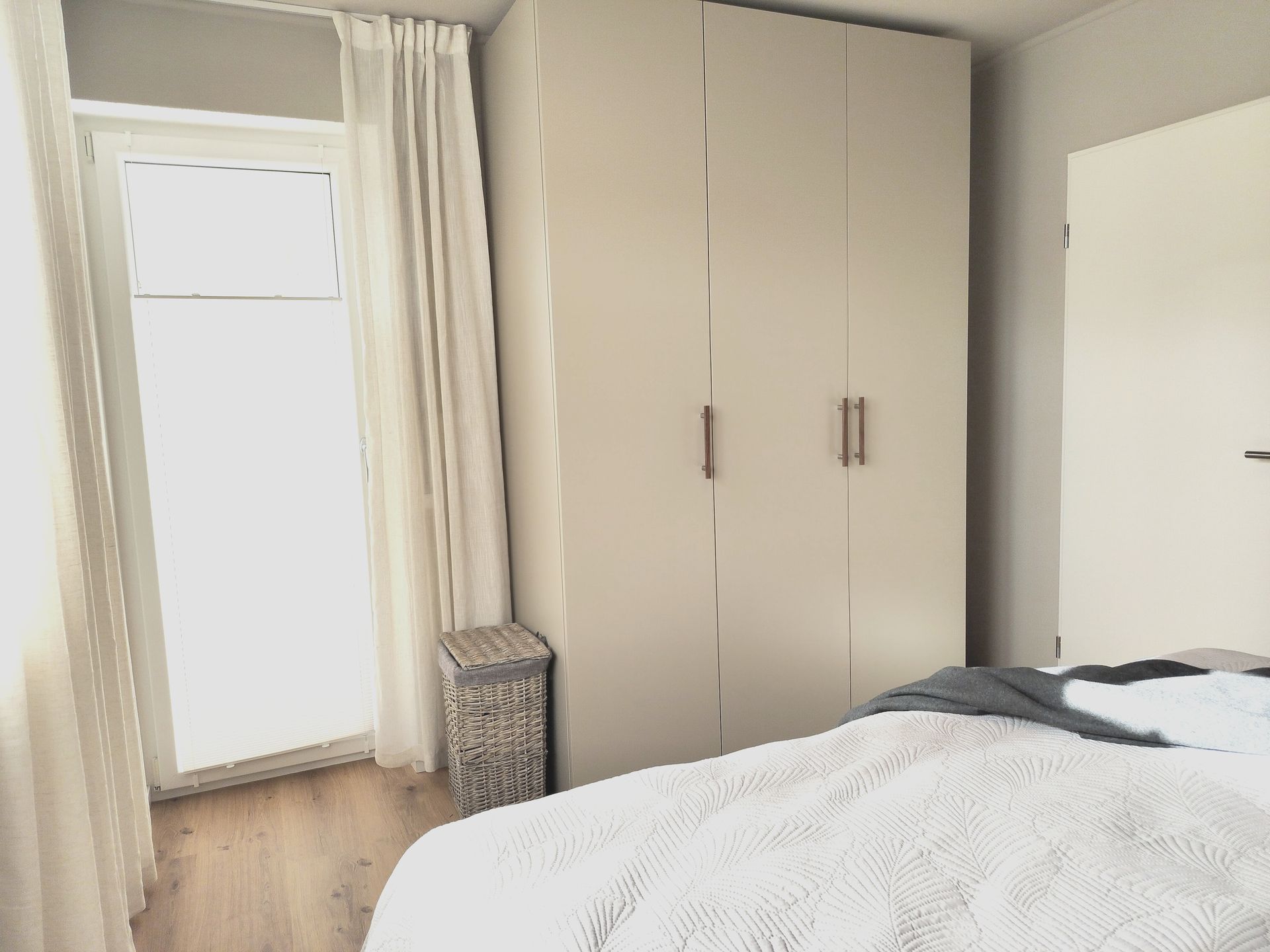 Helles Schlafzimmer mit Bett, hohem Kleiderschrank, Fenster mit Vorhängen und Weidenkorb auf Holzboden.