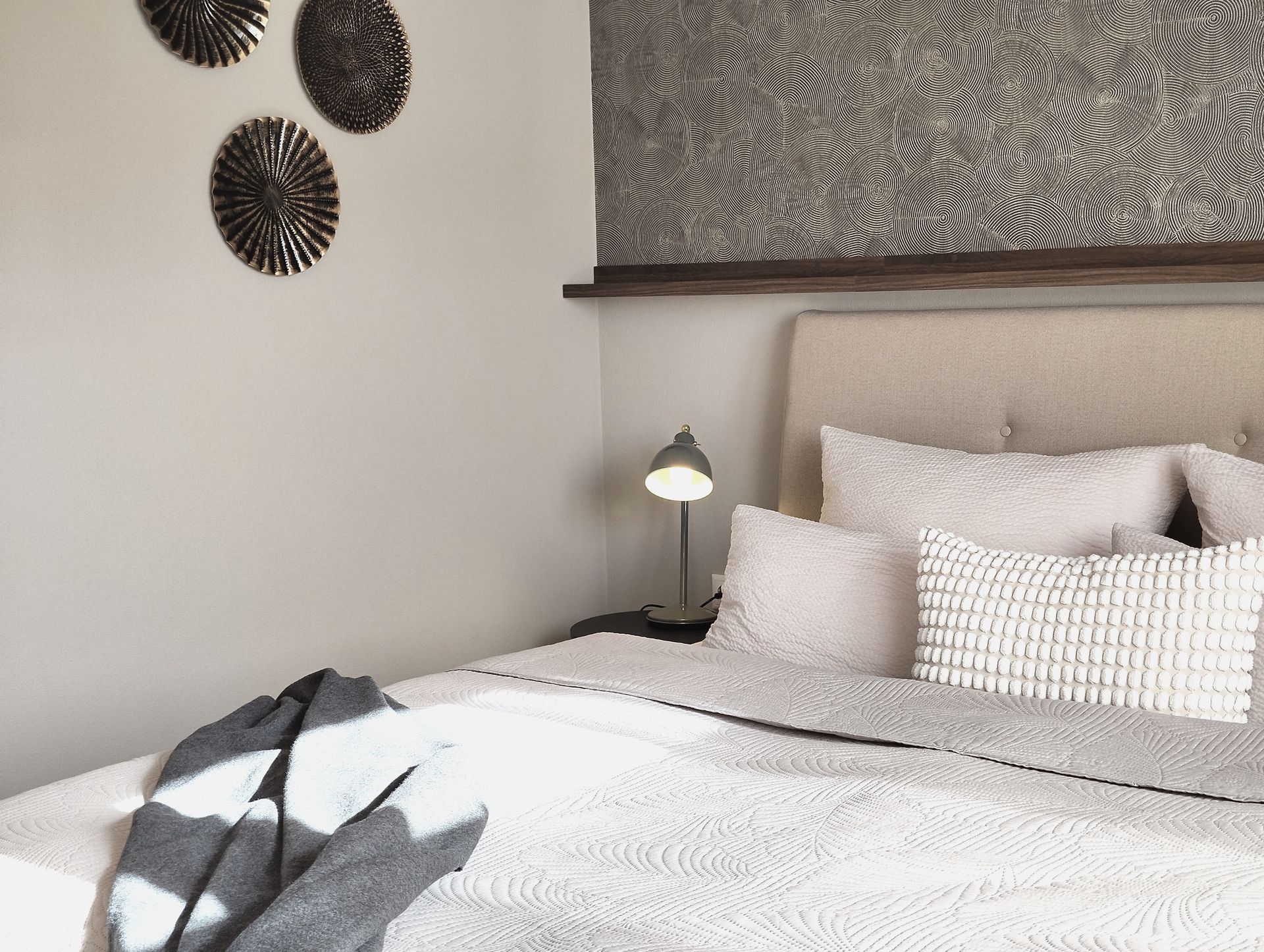 Ordentliches Schlafzimmer mit weißer Bettwäsche, Nachttischlampe und gemusterten Wandbildern in sanften, neutralen Tönen.