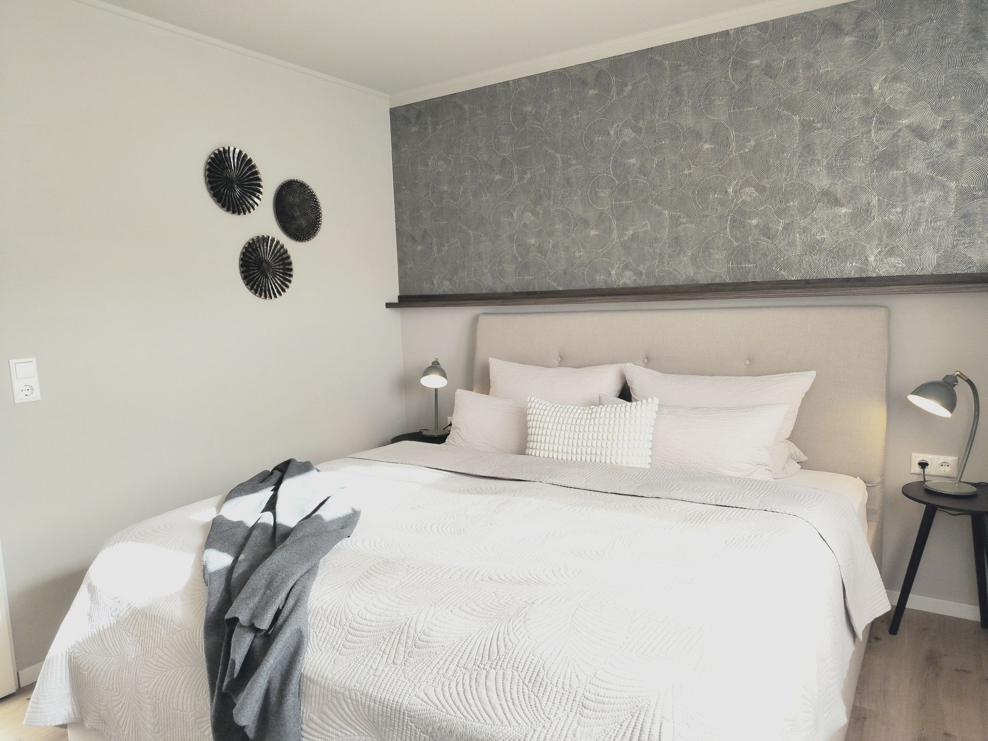 Helles Schlafzimmer mit ordentlich gemachtem Bett, grauer, strukturierter Kopfteilwand und kleinen Nachttischlampen.