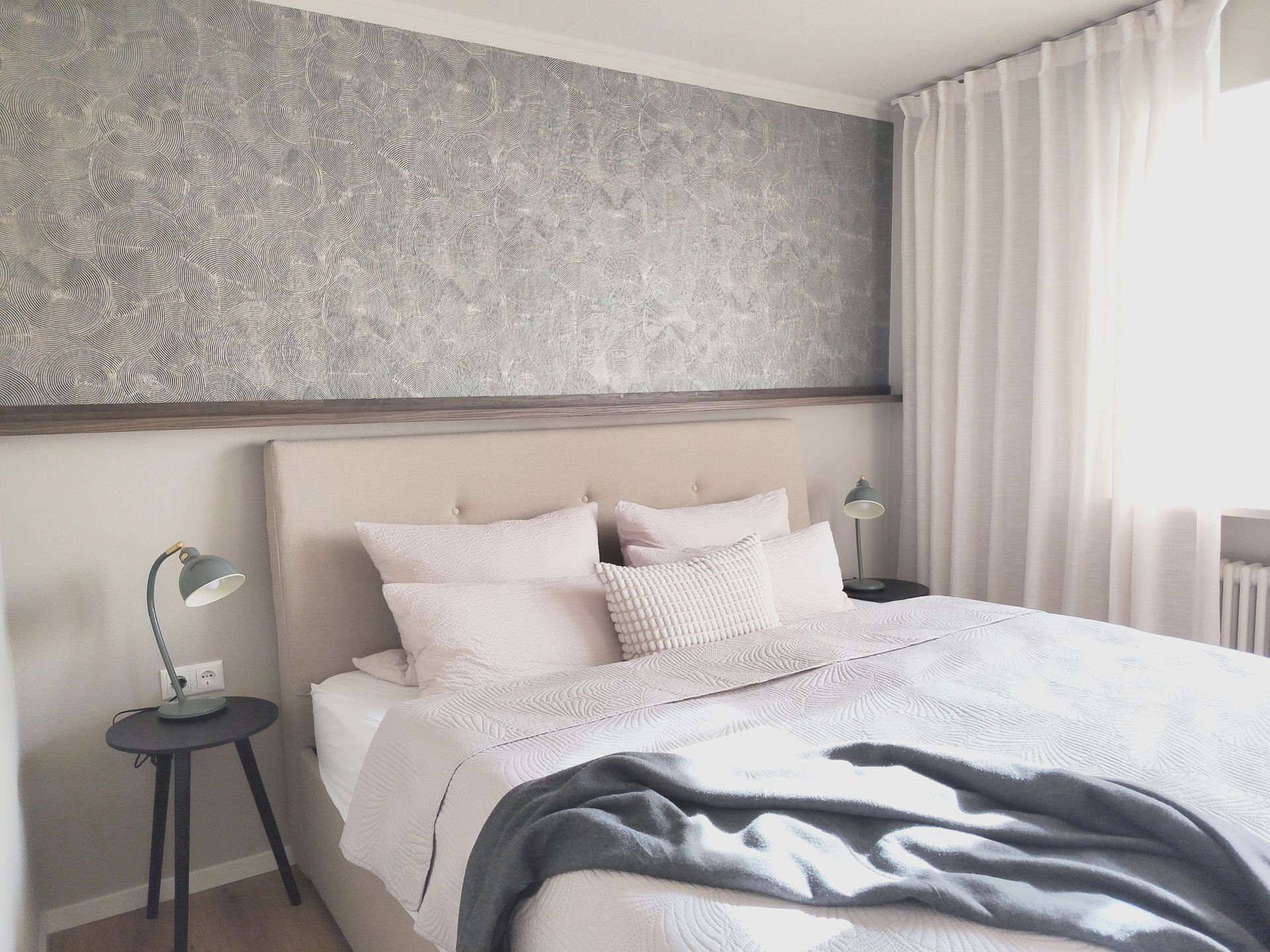 Ein ordentlich gemachtes Bett mit weißer Bettwäsche, grauer Tagesdecke, Nachttisch und strukturierter Wand in einem hellen Schlafzimmer