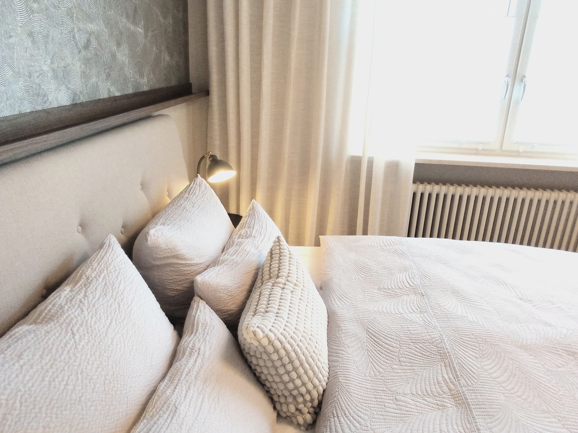 Gemütliche Schlafzimmerecke mit Bett, cremefarbenen Kissen, Lampe, Vorhängen und Sonnenlicht durch ein Fenster