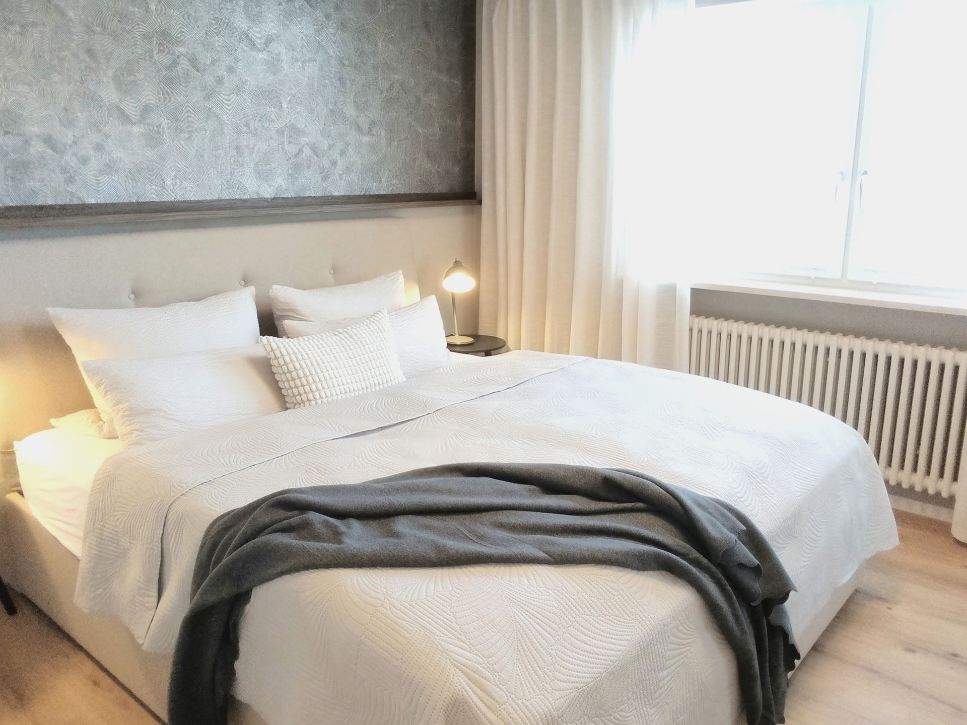 Helles Schlafzimmer mit ordentlich gemachtem weißen Bett, grauer Tagesdecke, Nachttischlampe und großem Fenster mit Vorhängen.