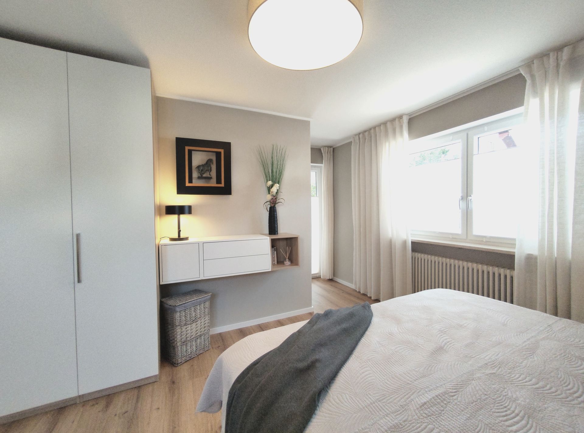 Helles Schlafzimmer mit Bett, Kleiderschrank, Schreibtisch, Lampe und großem Fenster mit Vorhängen.