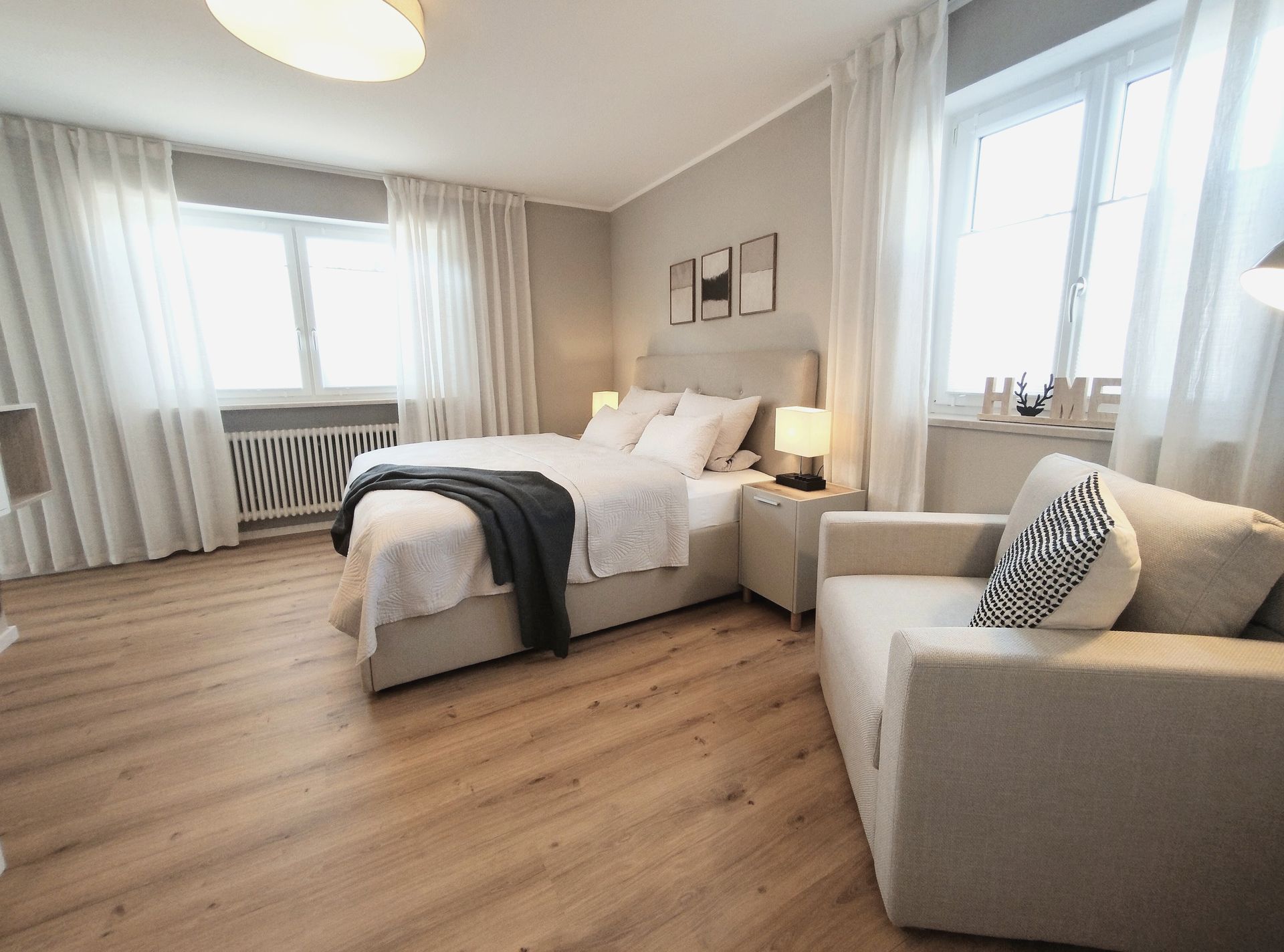 Helles Schlafzimmer mit weißem Bett, beigefarbenem Sessel, transparenten Vorhängen und Holzboden