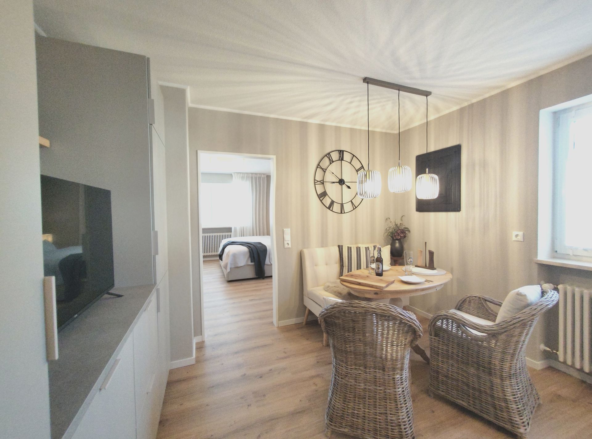 Heller Essbereich mit Korbstühlen, Holztisch, Wanduhr und offener Tür zu einem Schlafzimmer