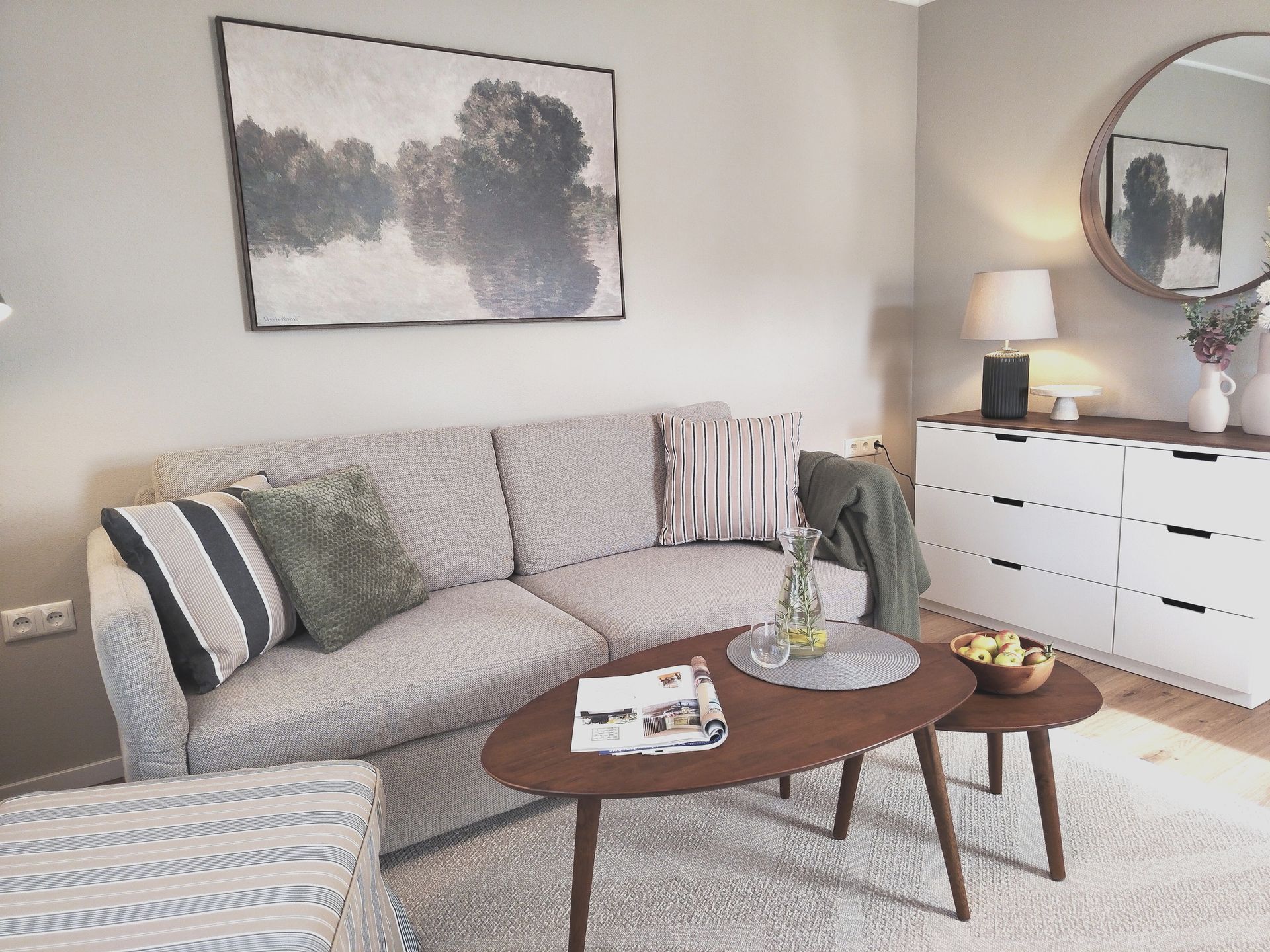 Helles Wohnzimmer mit grauem Sofa, Couchtischen, Kommode, Lampe und Wandbildern.