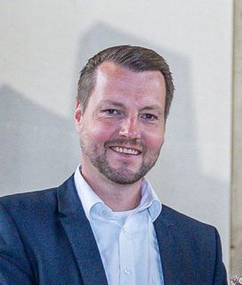 Eine lächelnde Person mit kurzem braunem Haar und Bart, die ein weißes Hemd mit Kragen und einen dunkelblauen Blazer trägt.