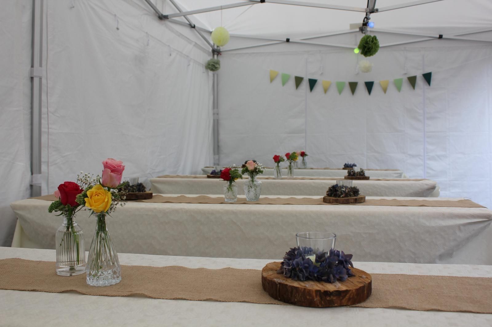 Tische in einem weißen Festzelt, dekoriert mit Juteläufern, kleinen Rosenvasen und Kerzenarrangements aus Holz.