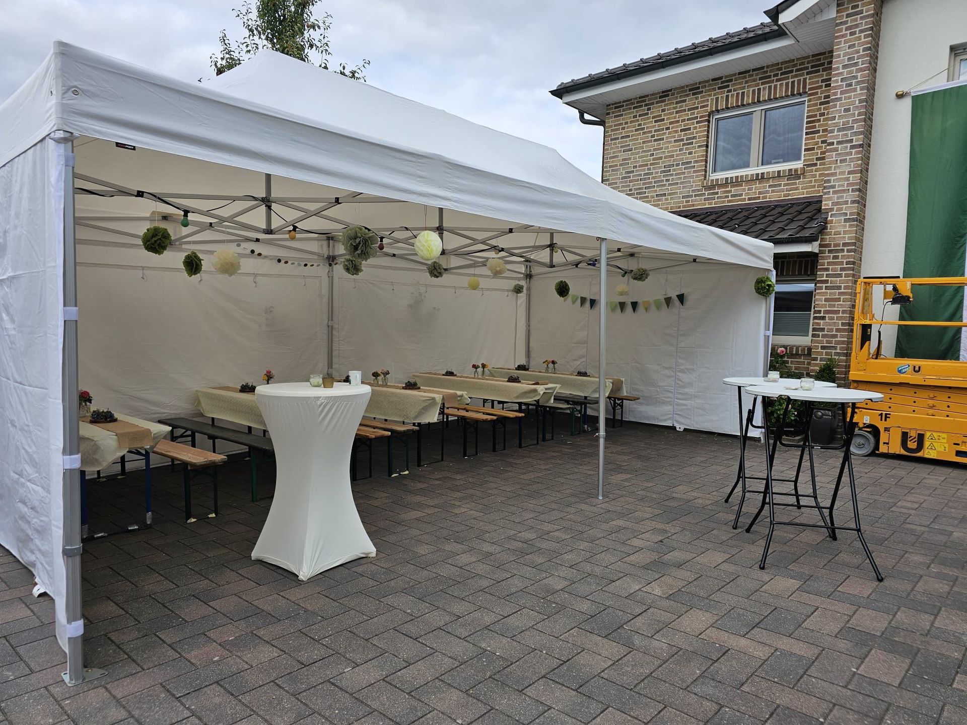Ein weißes Faltpavillon auf einer mit Ziegeln gepflasterten Terrasse, ausgestattet mit Tischen und Stühlen für eine Feier, daneben ein gelber Aufzug.