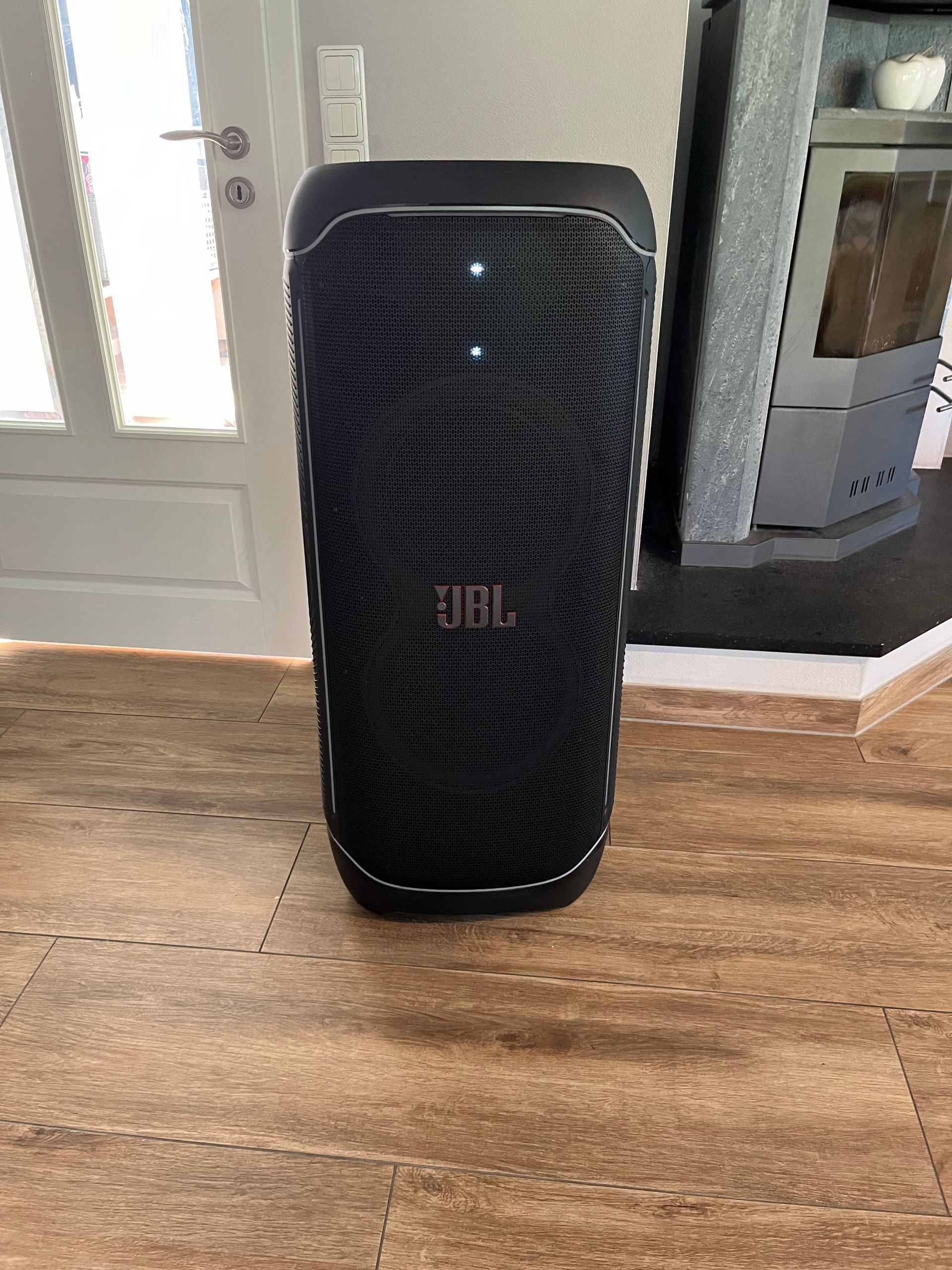 Ein schwarzer tragbarer JBL PartyBox-Lautsprecher steht auf einem Holzboden in einem Wohnraum.