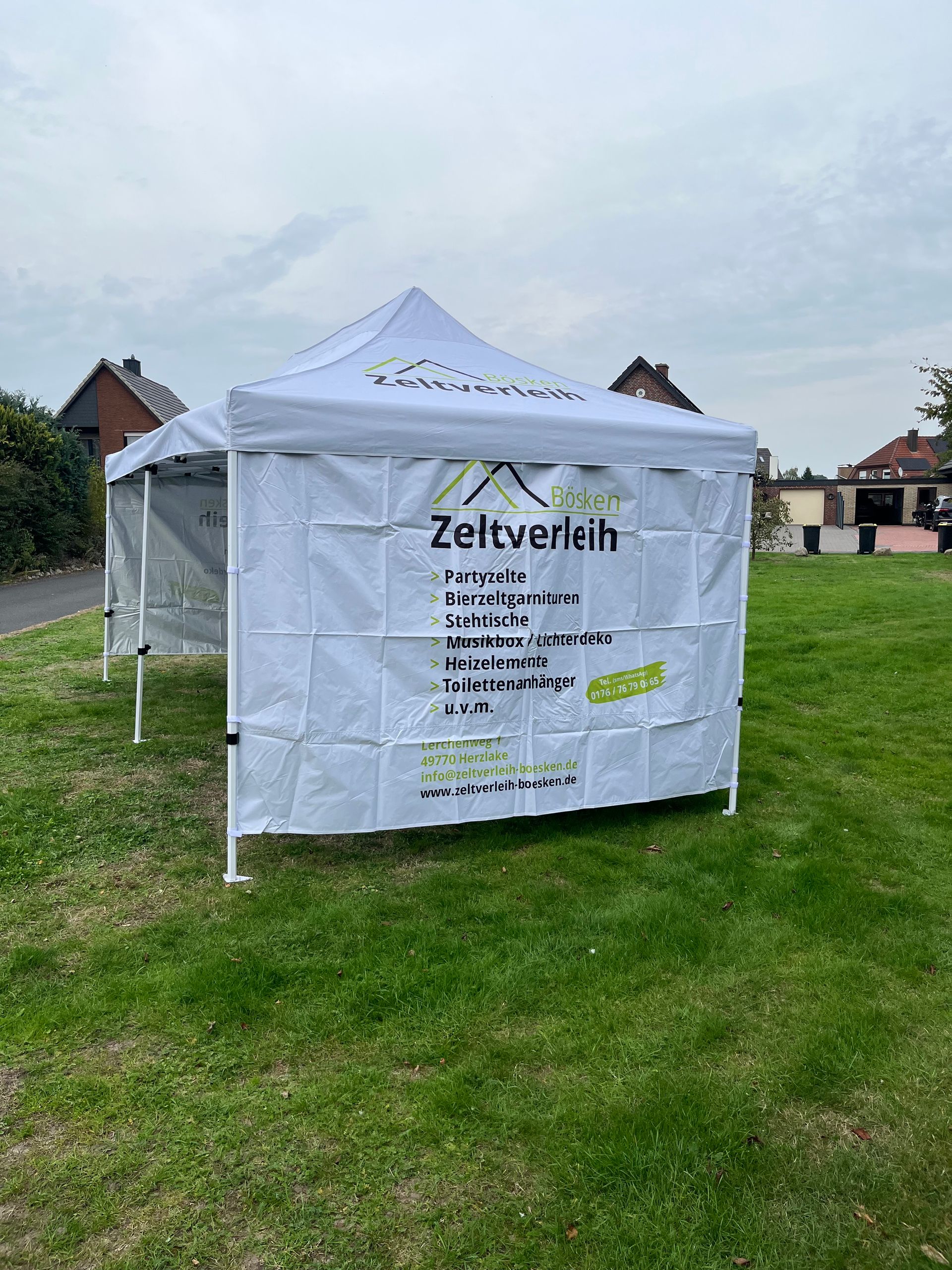 Ein weiß-graues Partyzelt mit verglasten Seitenwänden, aufgestellt auf einer Rasenfläche neben einer gemauerten Terrasse.