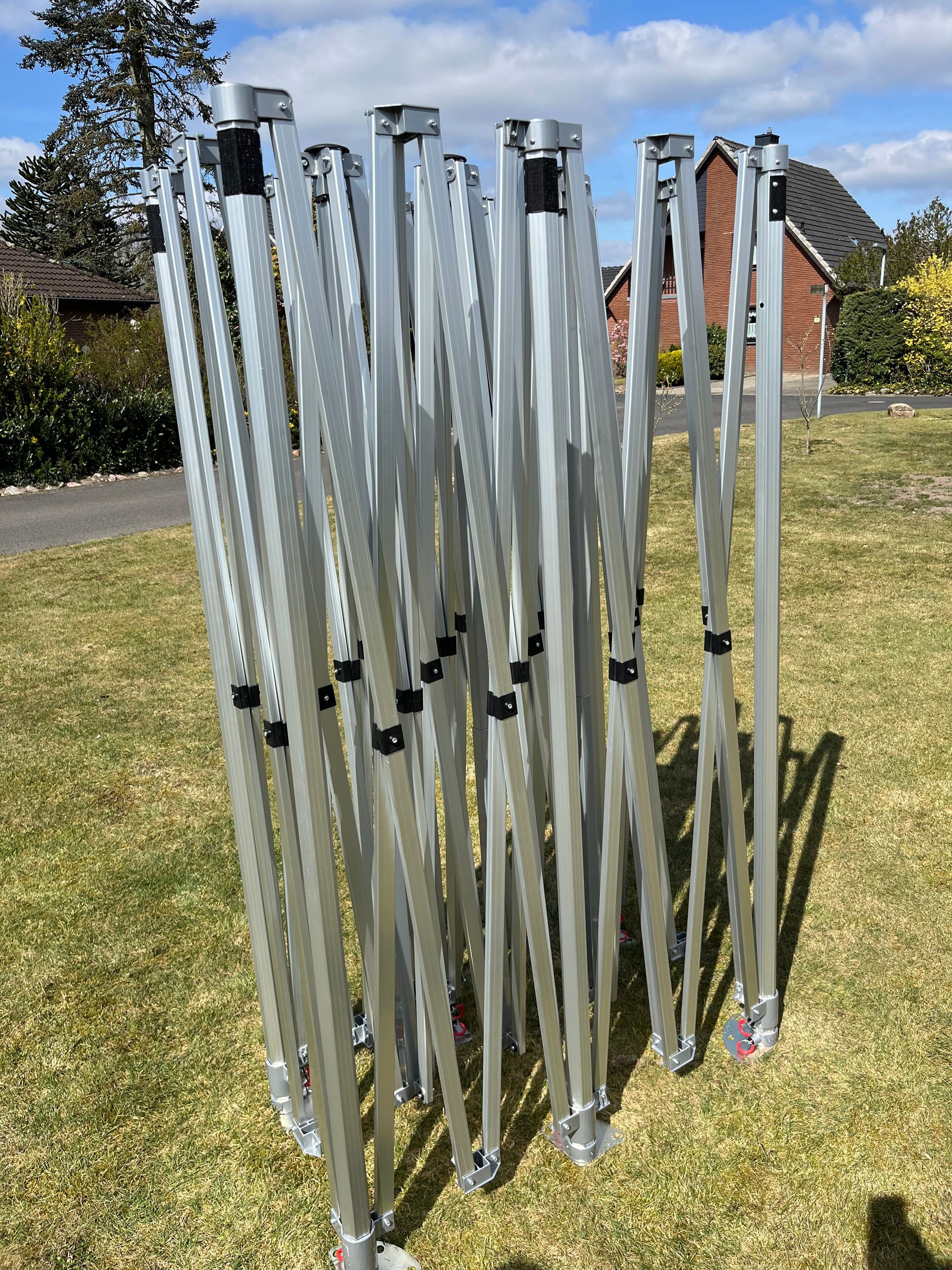 Ein teilweise zusammengebrochenes, silberfarbenes Metallgestell eines Faltpavillons steht auf einer Rasenfläche.