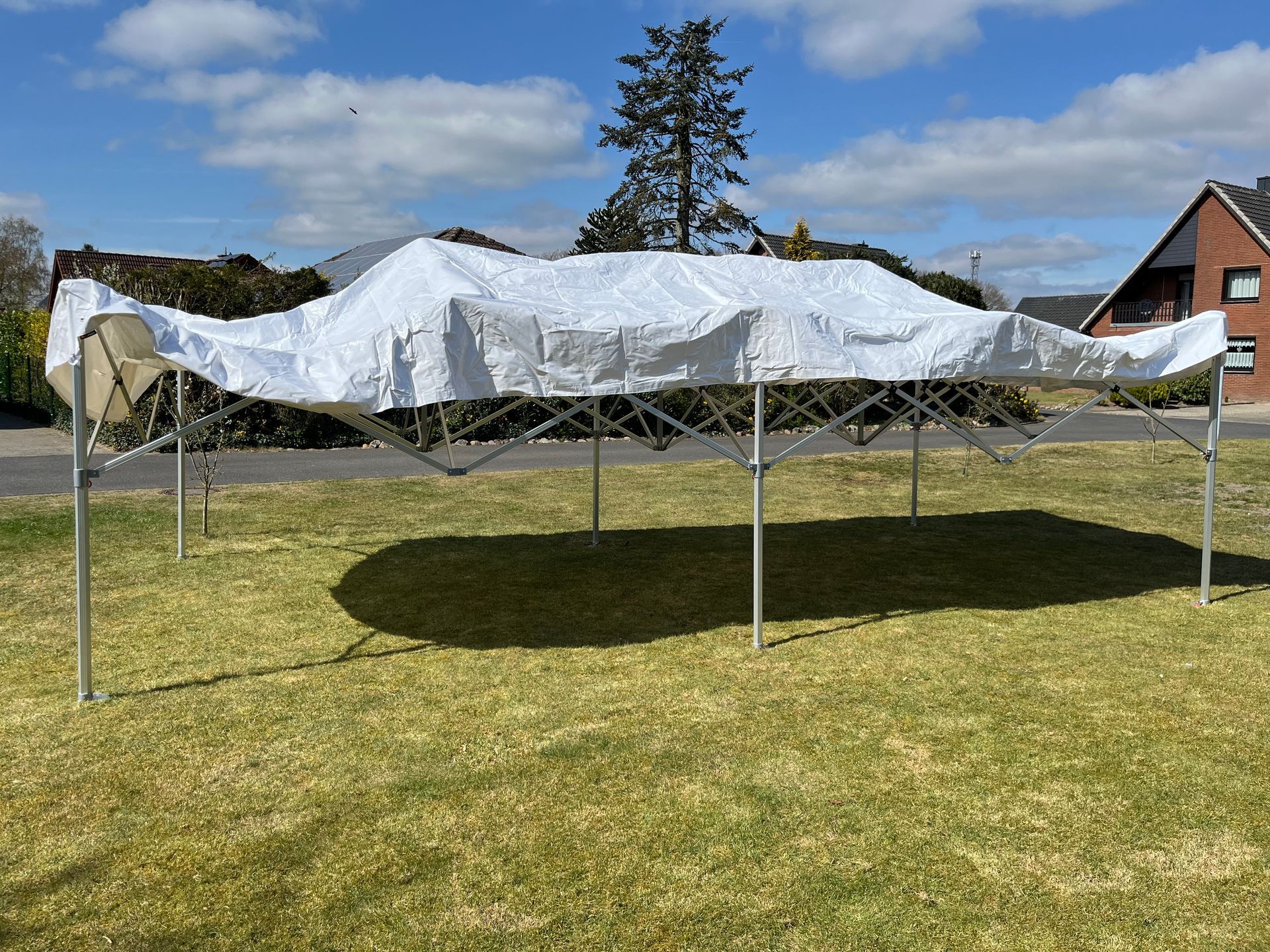 Ein weißes Pop-up-Zeltgestell mit einem drapierten, teilweise entfalteten weißen Baldachin steht auf einer Rasenfläche unter blauem Himmel.