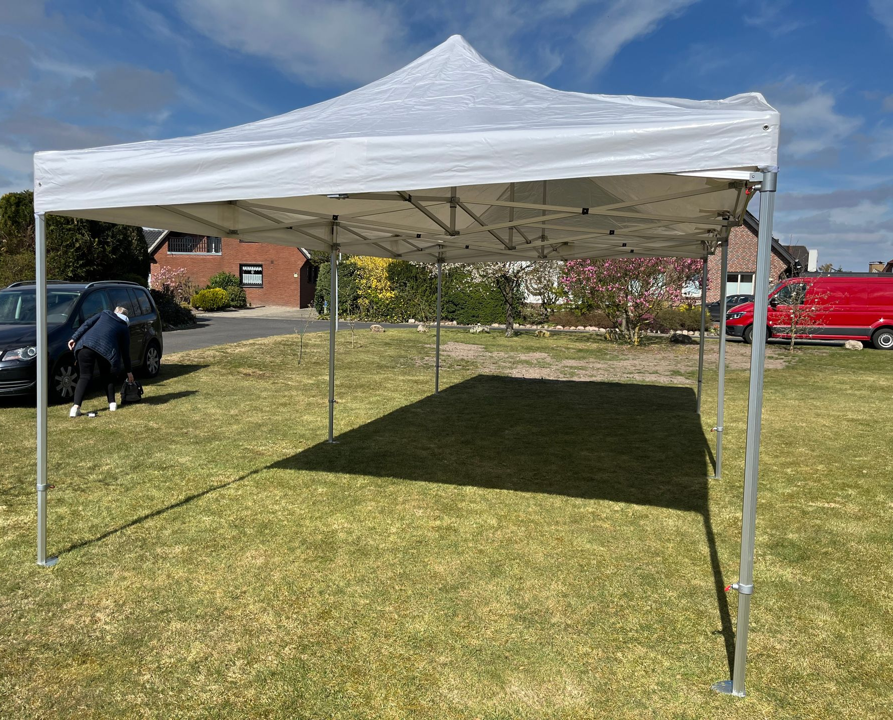 Auf einer sonnigen Rasenfläche steht ein weißes Faltpavillonzelt; im Hintergrund sind eine Person und geparkte Fahrzeuge zu sehen.