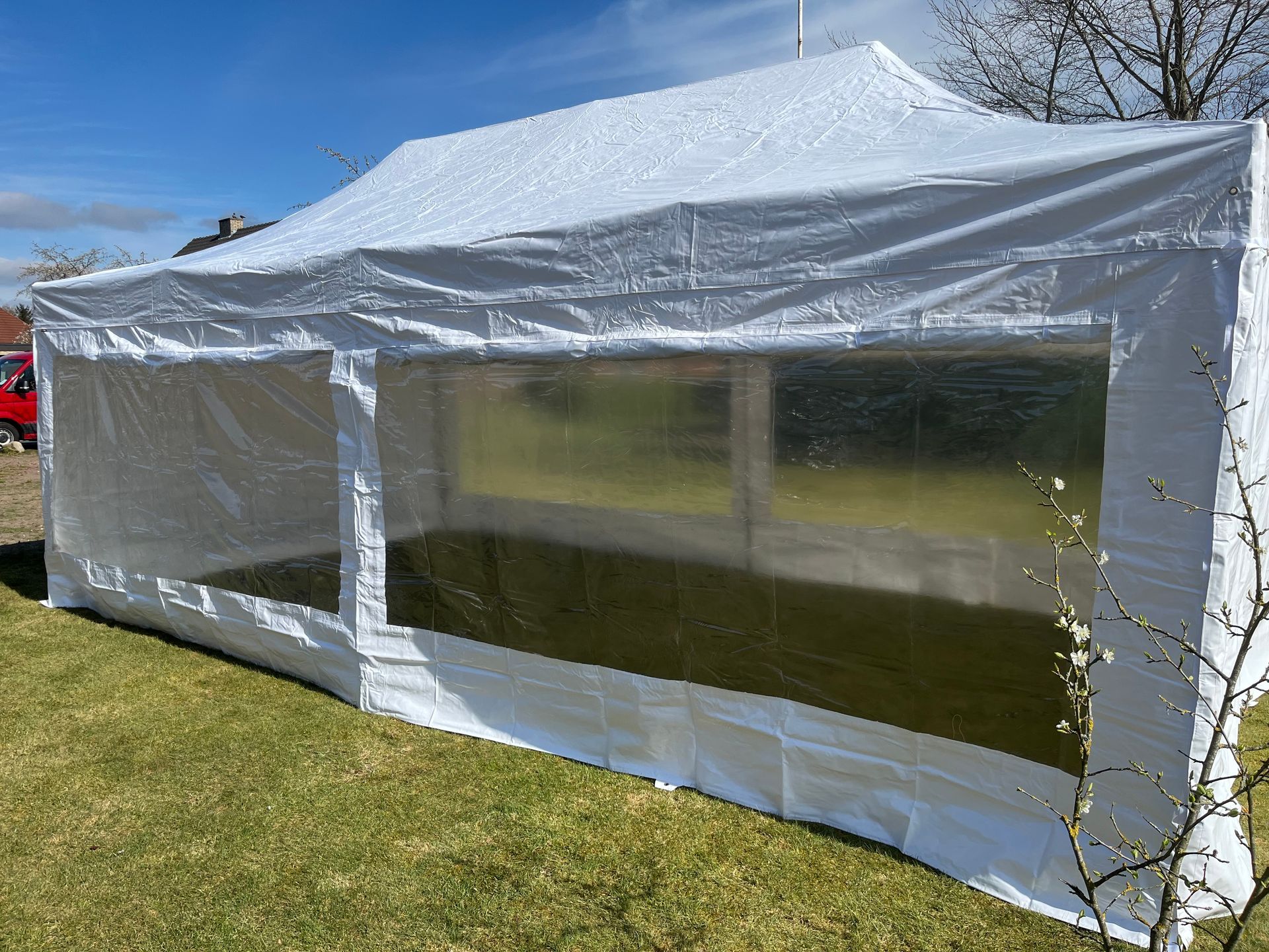 Ein weißes, rechteckiges Partyzelt mit großen, durchsichtigen Kunststofffenstern, aufgebaut auf einer Rasenfläche unter strahlend blauem Himmel.