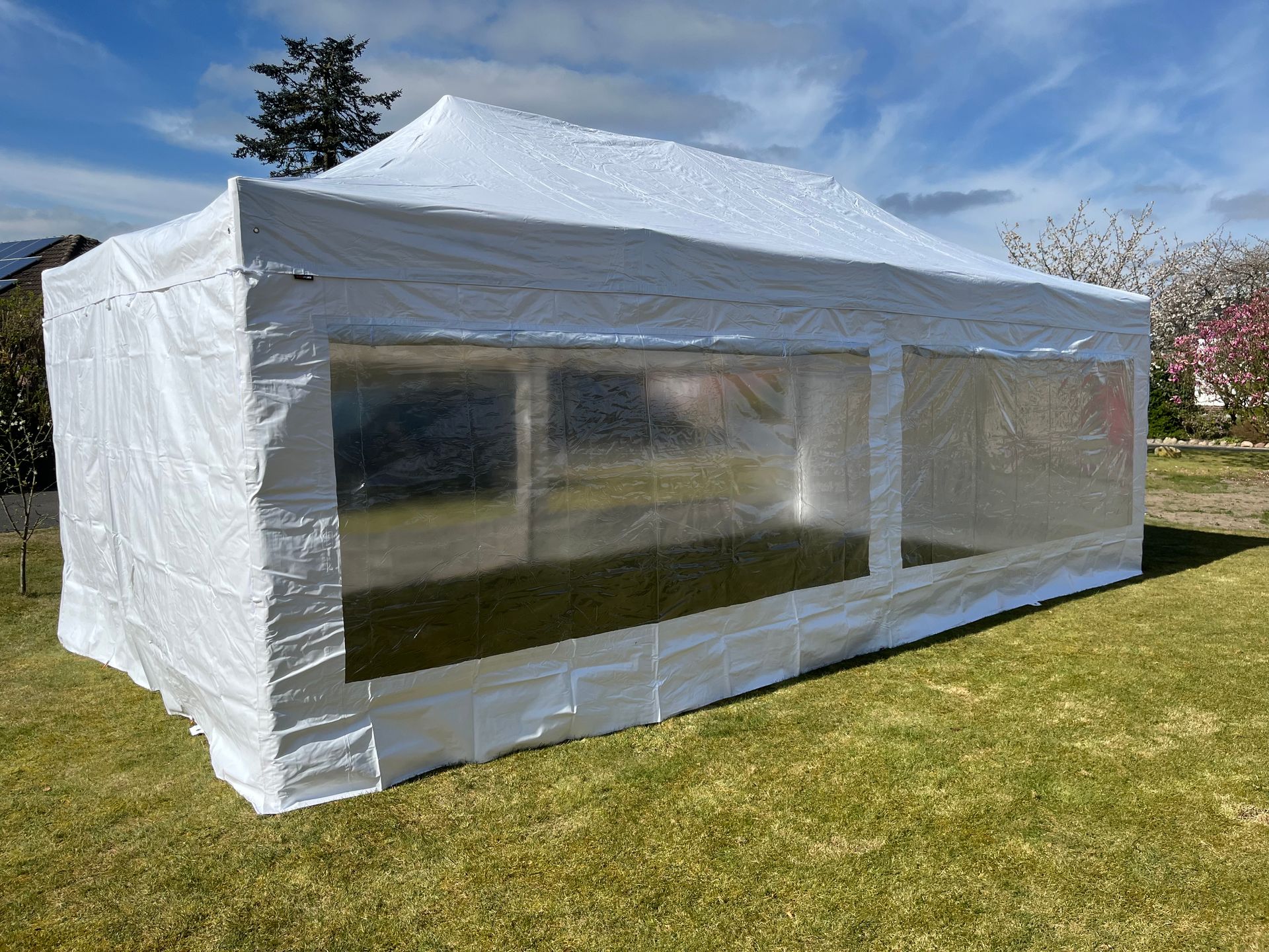 Ein großes, weißes, rechteckiges Veranstaltungszelt mit transparenten Fenstern, das auf einer Rasenfläche unter klarem, blauem Himmel steht.