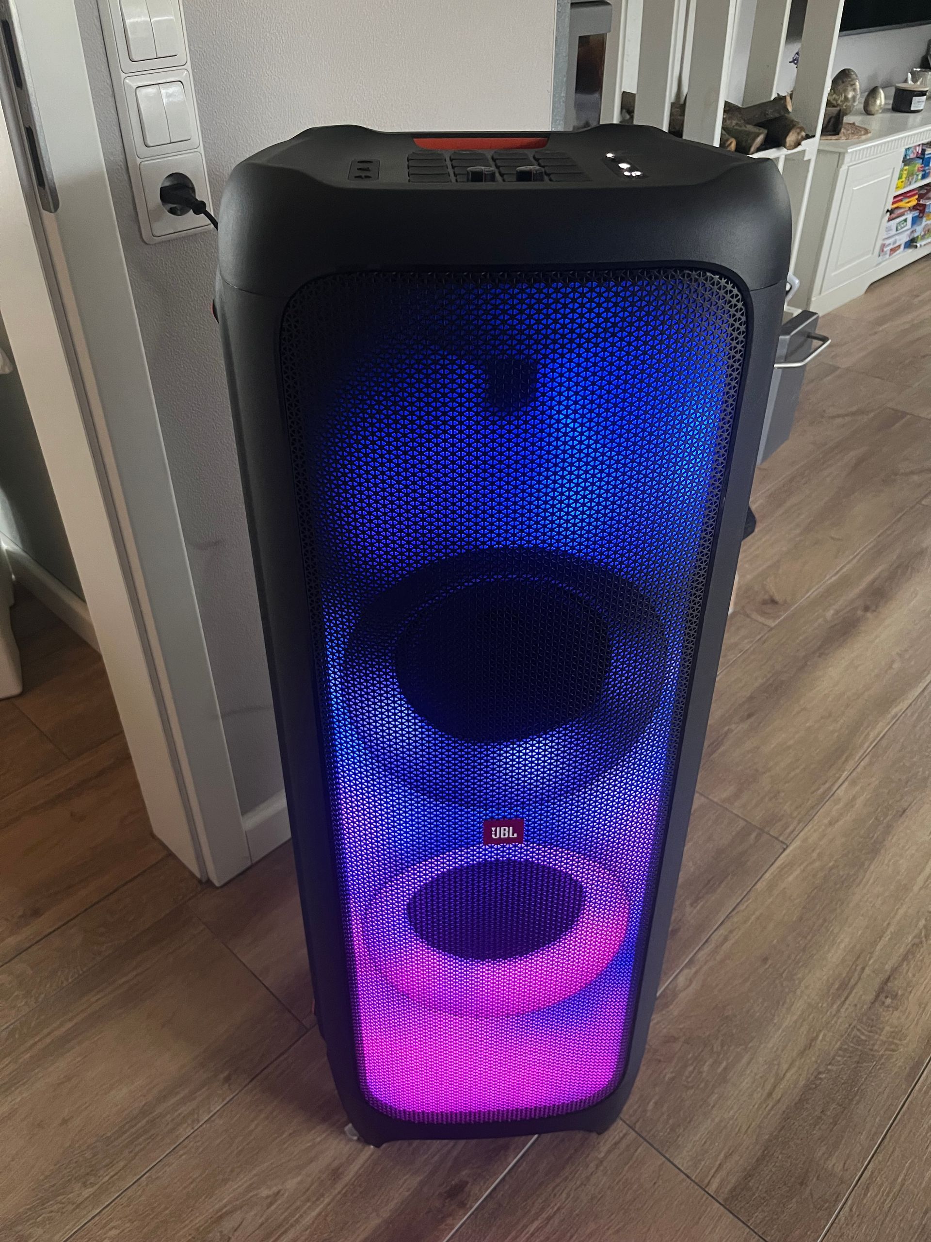 Ein hoher, schwarzer JBL PartyBox-Lautsprecher steht auf einem Holzboden und verfügt über eine Frontplatte, die in Blau und Rosa leuchtet.