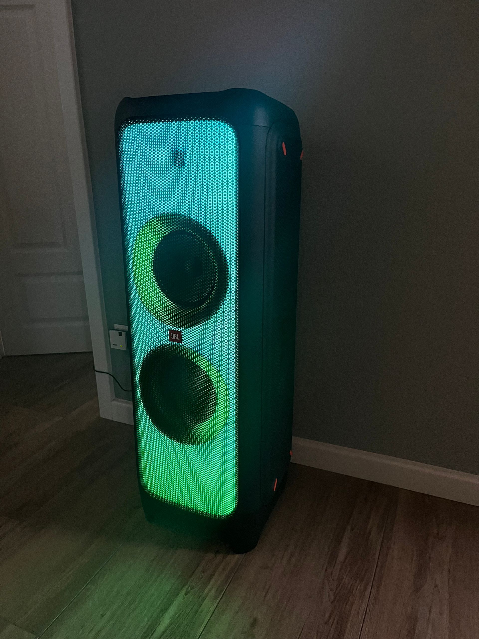 Ein hoher JBL PartyBox-Lautsprecher, der mit hellgrünen LED-Lichtmustern erstrahlt, steht auf einem Hartholzboden neben einer Wand.