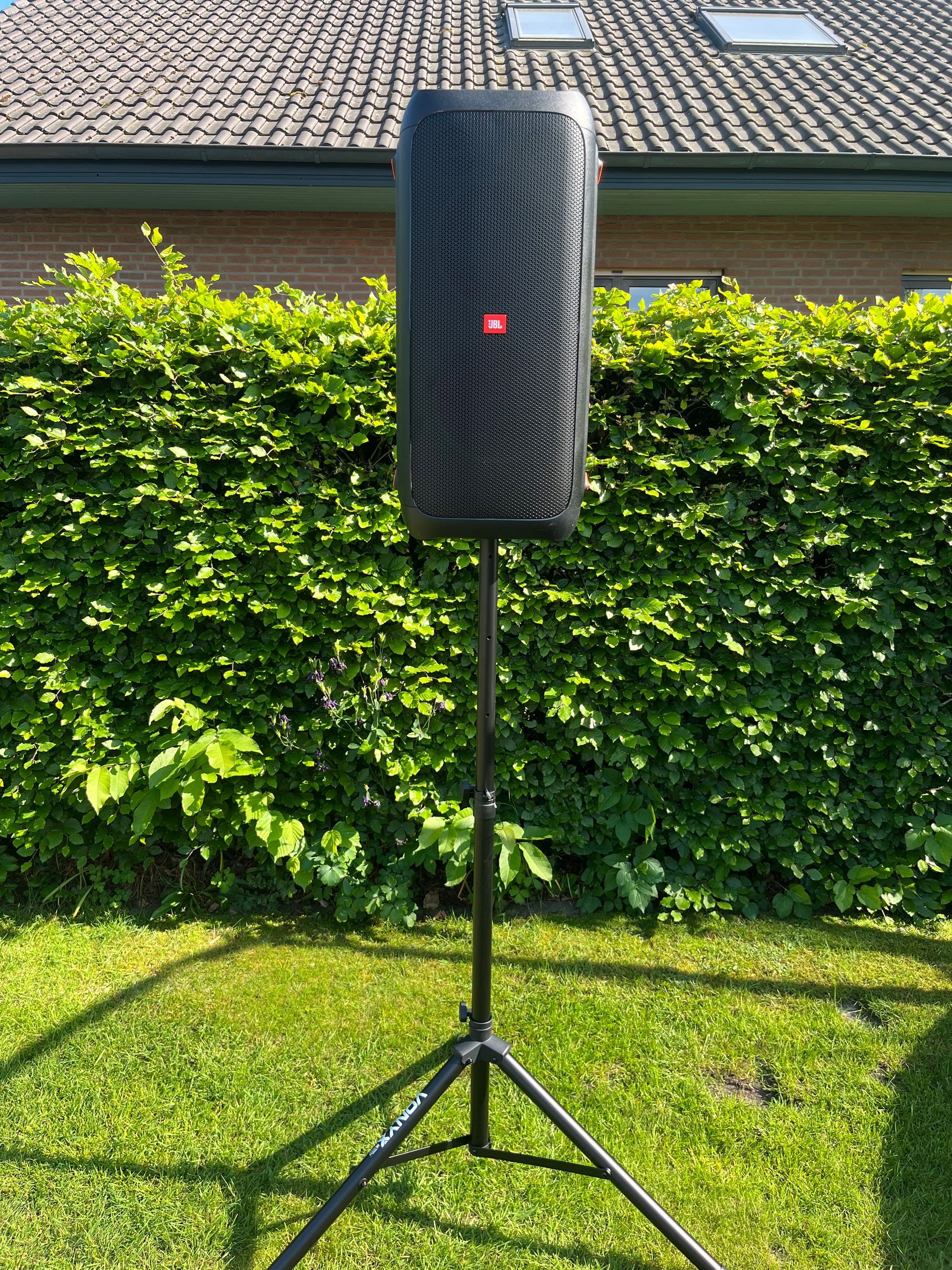 Ein schwarzer JBL-Lautsprecher, montiert auf einem Stativ, stand auf einer Rasenfläche vor einer grünen Hecke und einem Backsteinhaus.