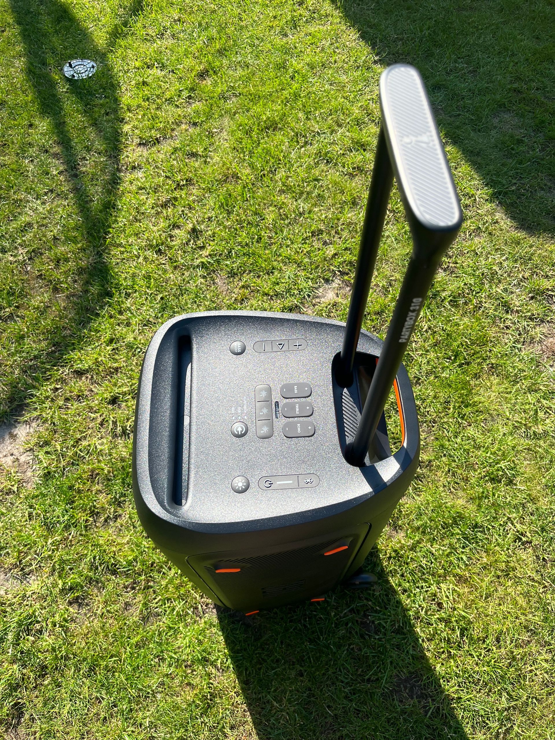 Ein schwarzer, tragbarer Lautsprecher mit einem ausziehbaren, kofferartigen Griff steht auf grünem Gras.