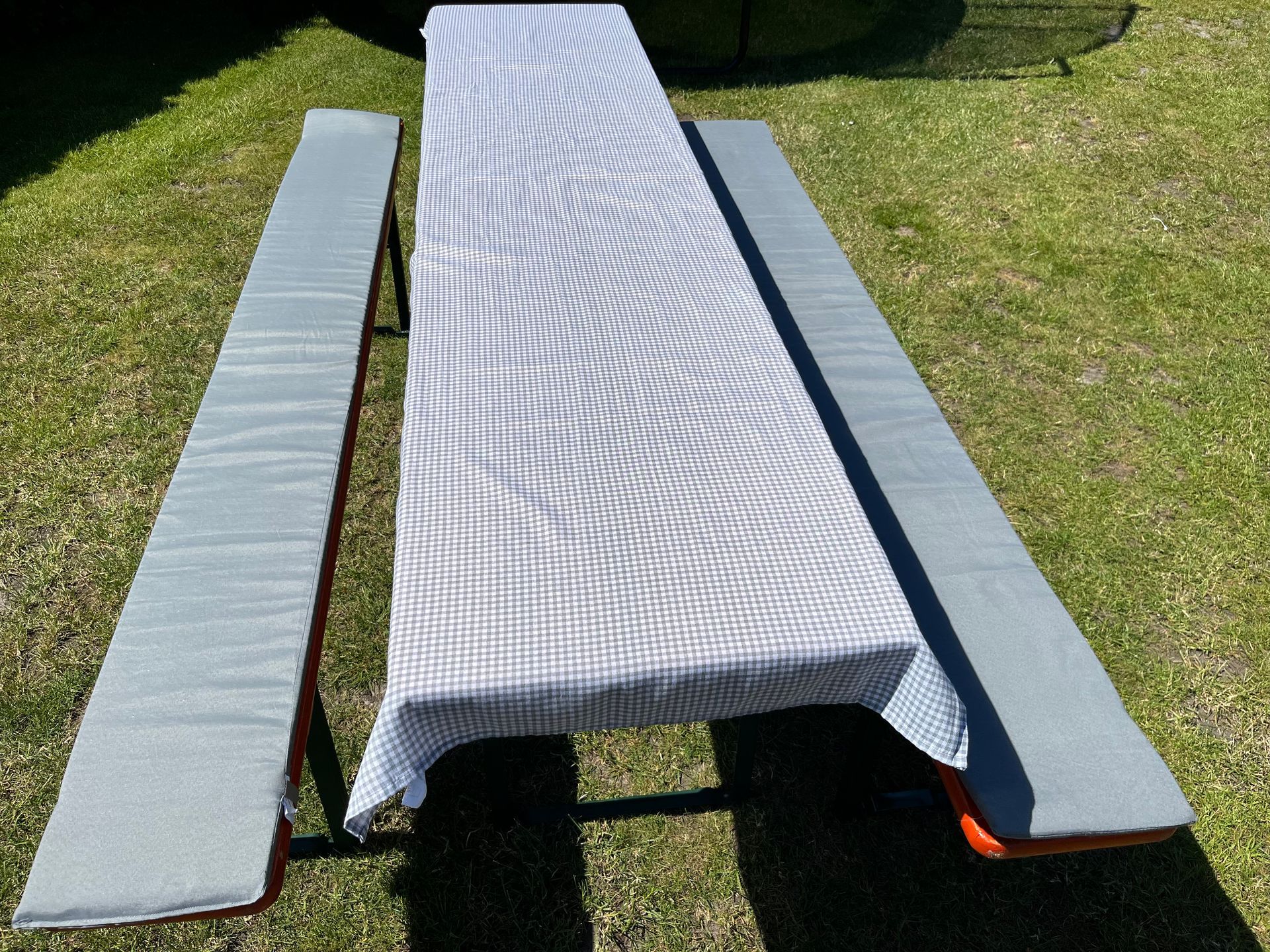 Ein langer Biergartentisch mit blau-weiß karierter Tischdecke und zwei grauen Bankkissen auf einer Rasenfläche.
