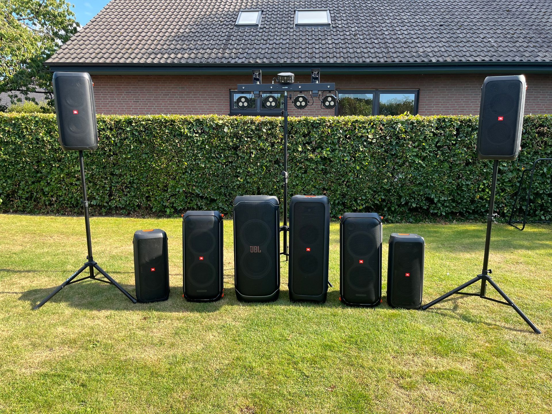 Eine Reihe schwarzer JBL-Lautsprecher auf Rasenflächen, zwei Lautsprecher auf Ständern und im Hintergrund eine Lichtleiste.