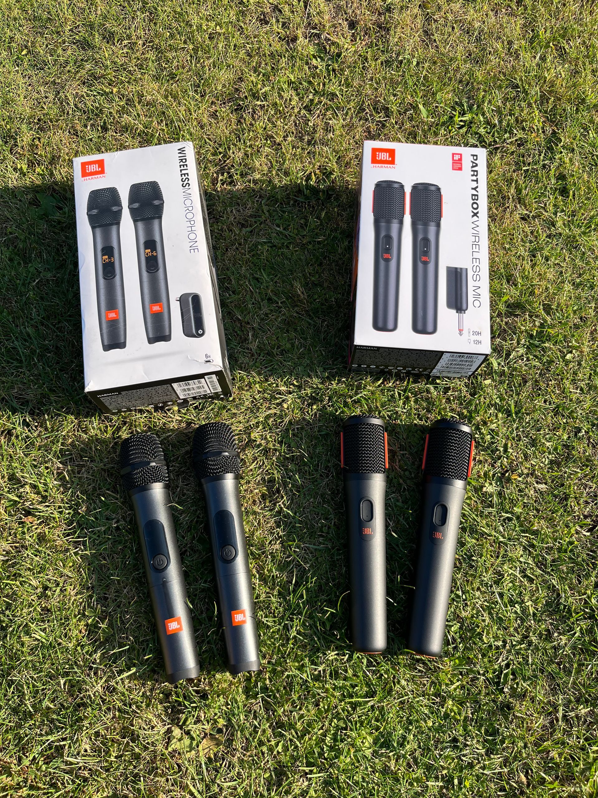 Zwei Paare drahtloser JBL-Mikrofone, jeweils mit zugehöriger Verpackung, sind vor einem grünen Grashintergrund angeordnet.