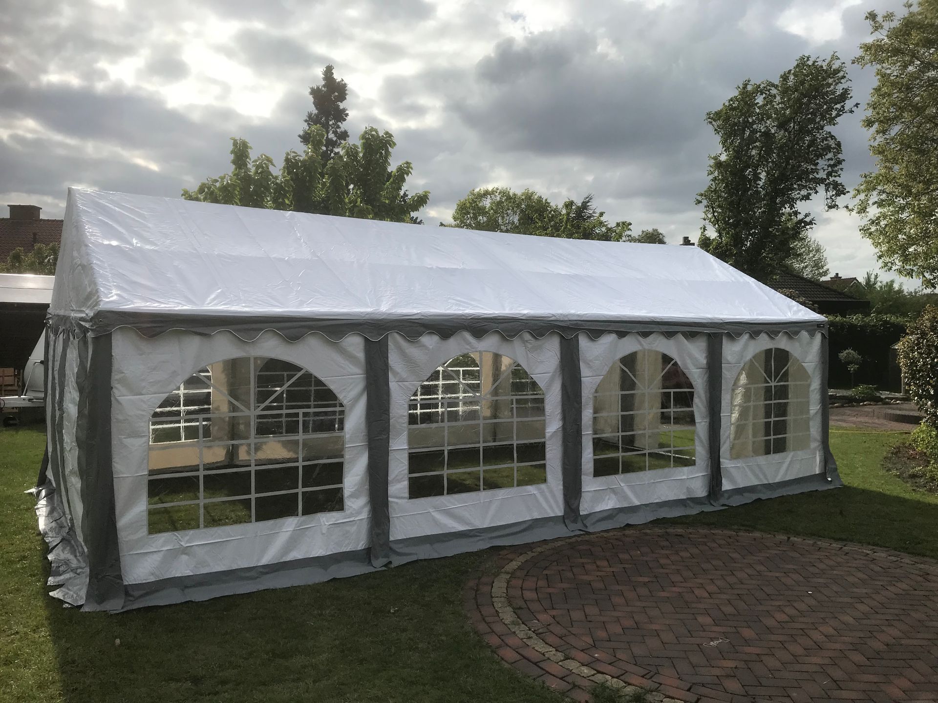 Ein großes weißes rechteckiges Partyzelt mit bogenförmigen transparenten Fenstern, aufgebaut auf einer Rasenfläche neben einer gemauerten Terrasse.