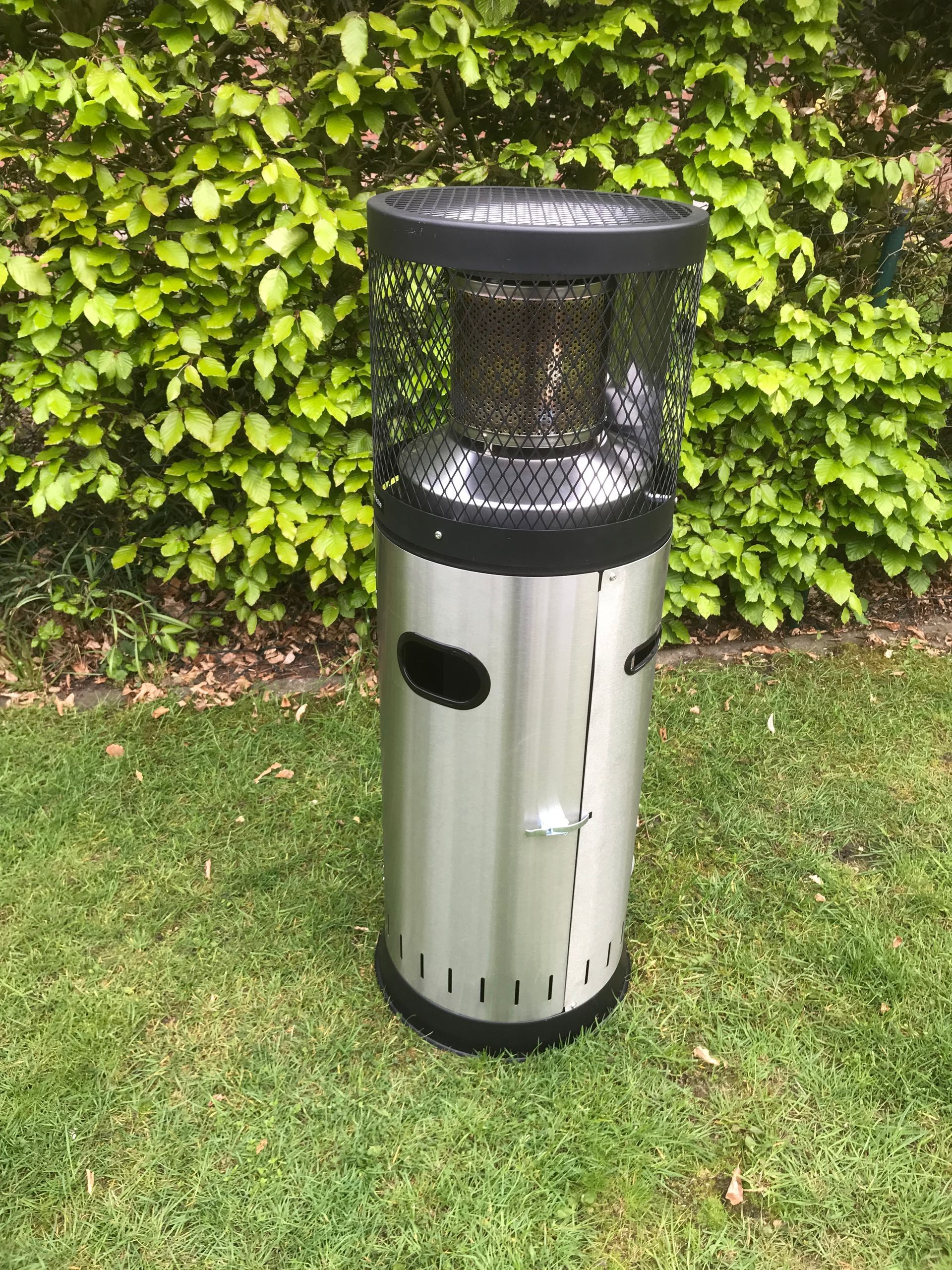 Ein Terrassenheizstrahler aus Edelstahl mit einem schützenden schwarzen Drahtgitter steht auf einer Rasenfläche vor grünen Büschen.