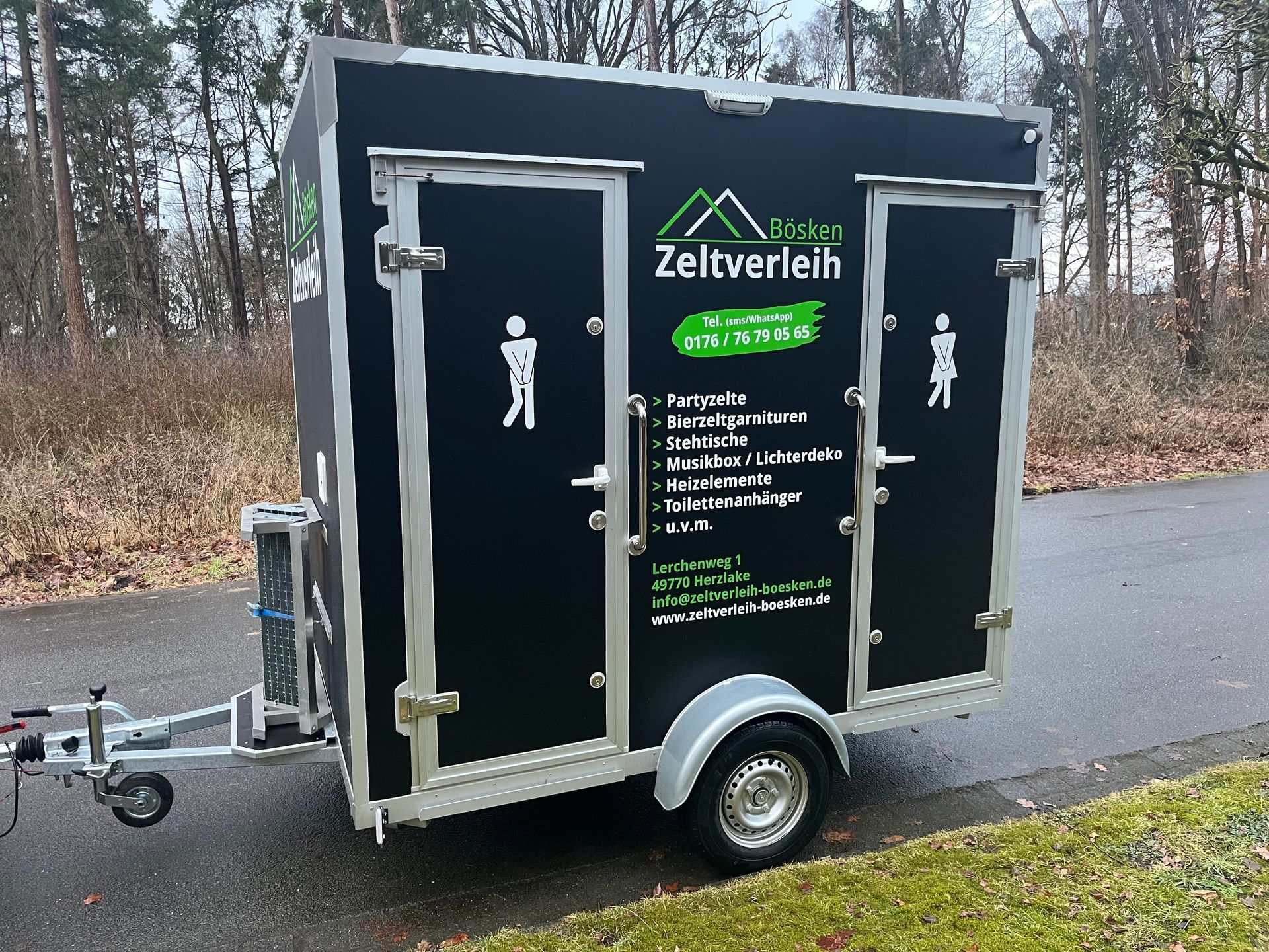 Ein schwarzer, mobiler Toilettenwagen mit zwei Türen steht auf einer Straße; er ist mit weißer Schrift und Symbolen für Herren- und Damentoiletten versehen.