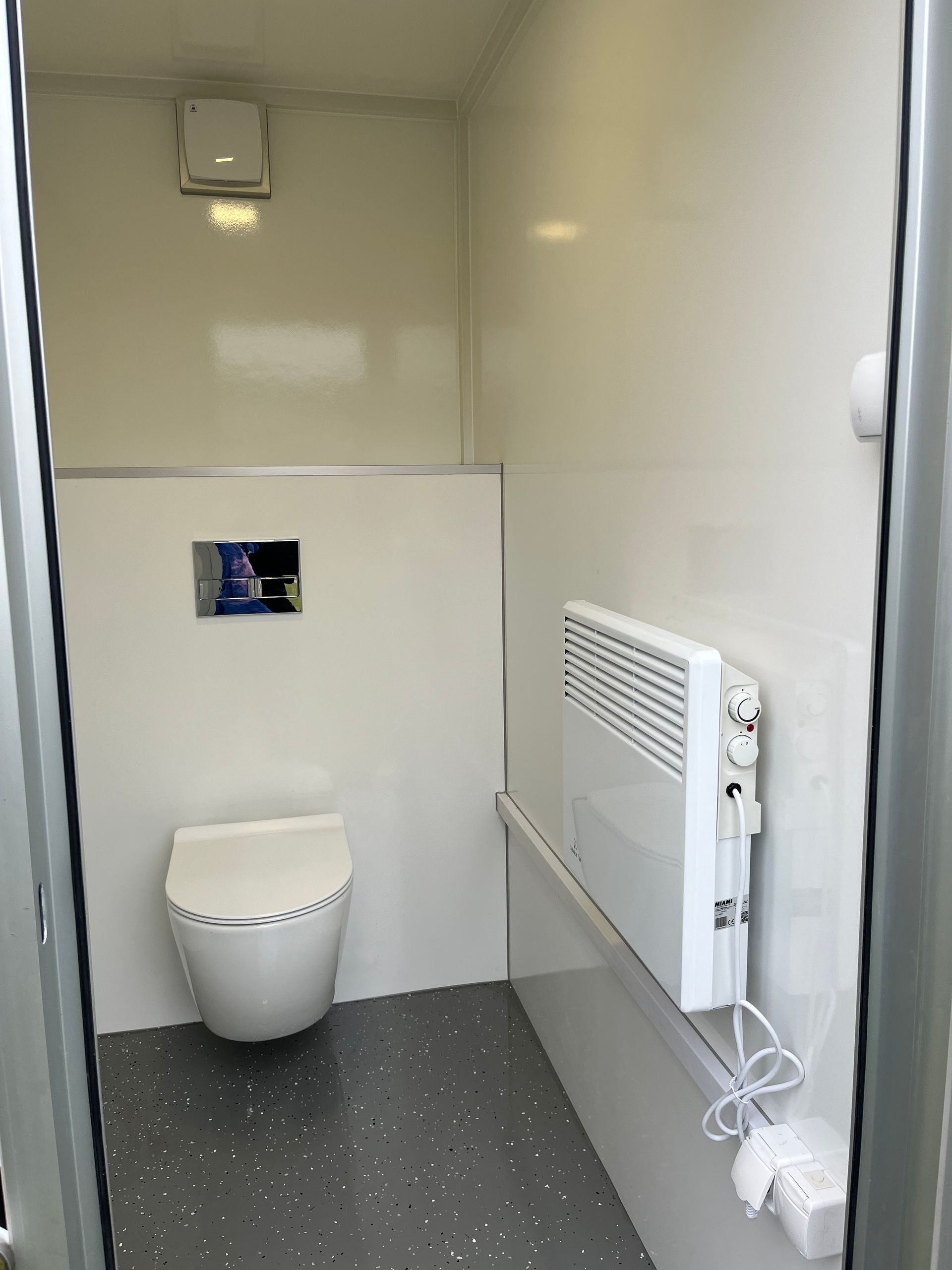 Eine kleine, weiße, moderne barrierefreie Toilettenkabine mit wandmontierter Toilette und elektrischer Wandheizung.