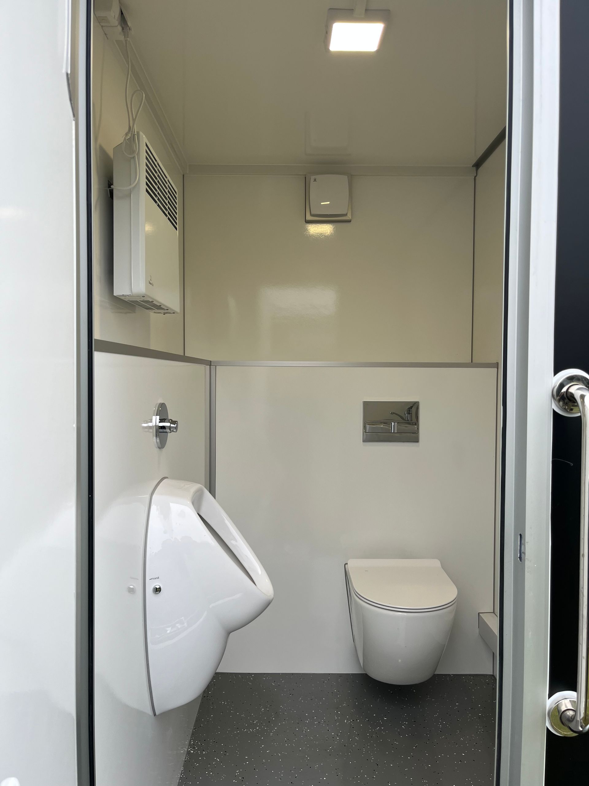 Eine kleine, saubere, weiße Modultoilette mit wandmontiertem Urinal, Toilette, Deckenleuchte und grau gesprenkeltem Boden.