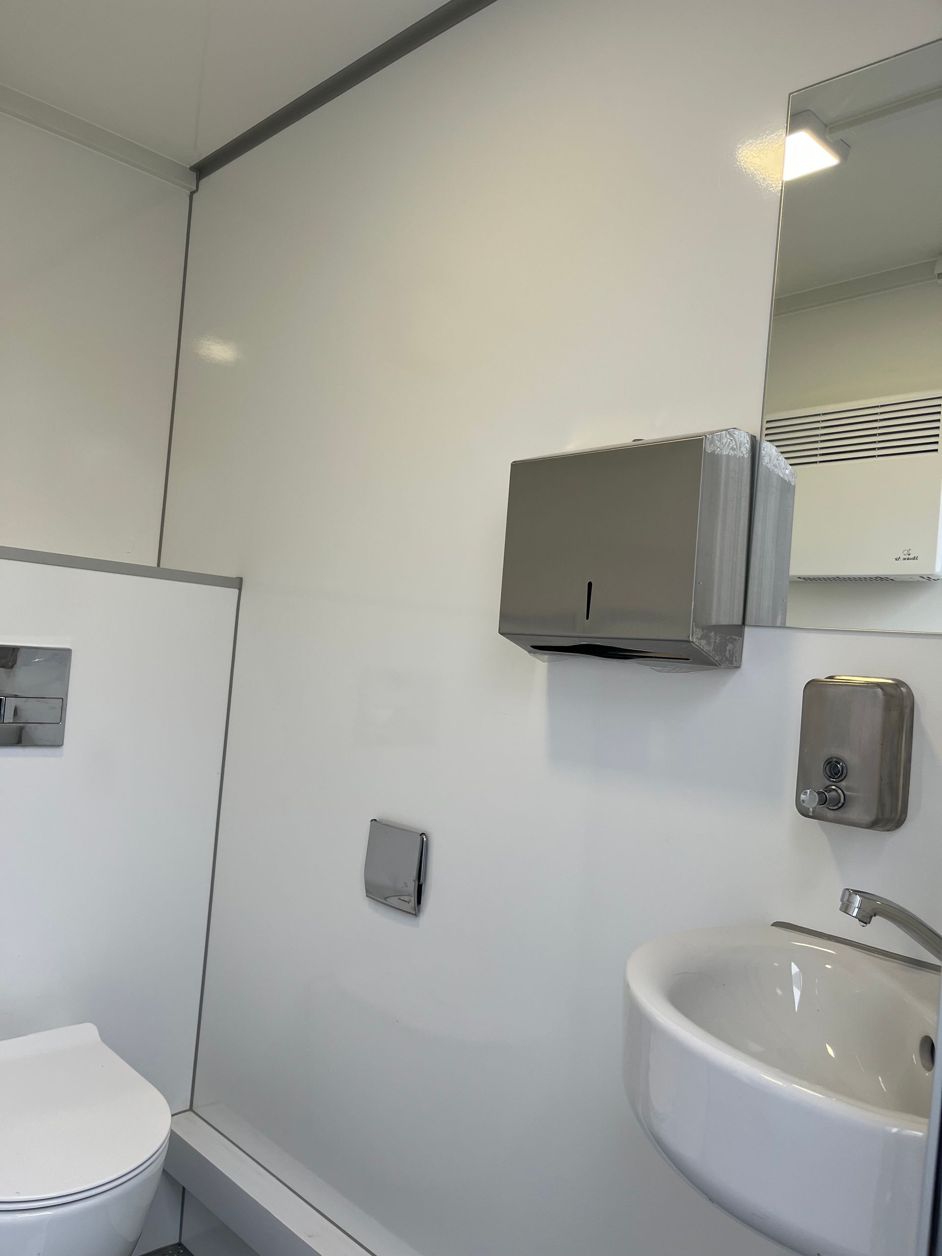 Ein helles, weißes Badezimmer mit Toilette, Waschbecken, einem grauen Papierhandtuchspender und einem an der Wand montierten Seifenspender.