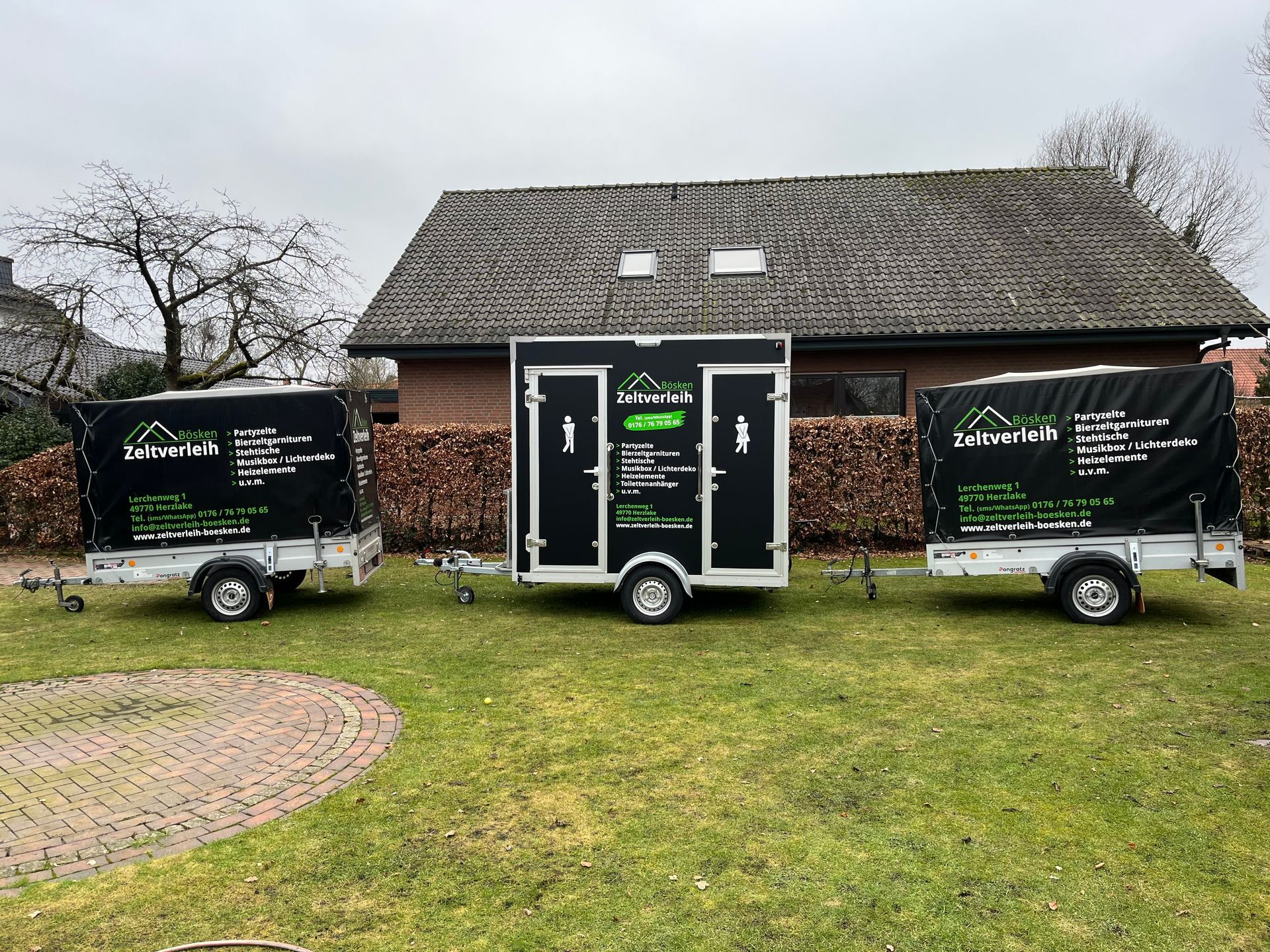 Drei schwarze, mobile Toilettenwagen standen auf einer Rasenfläche vor einem Haus mit Schindeldach.