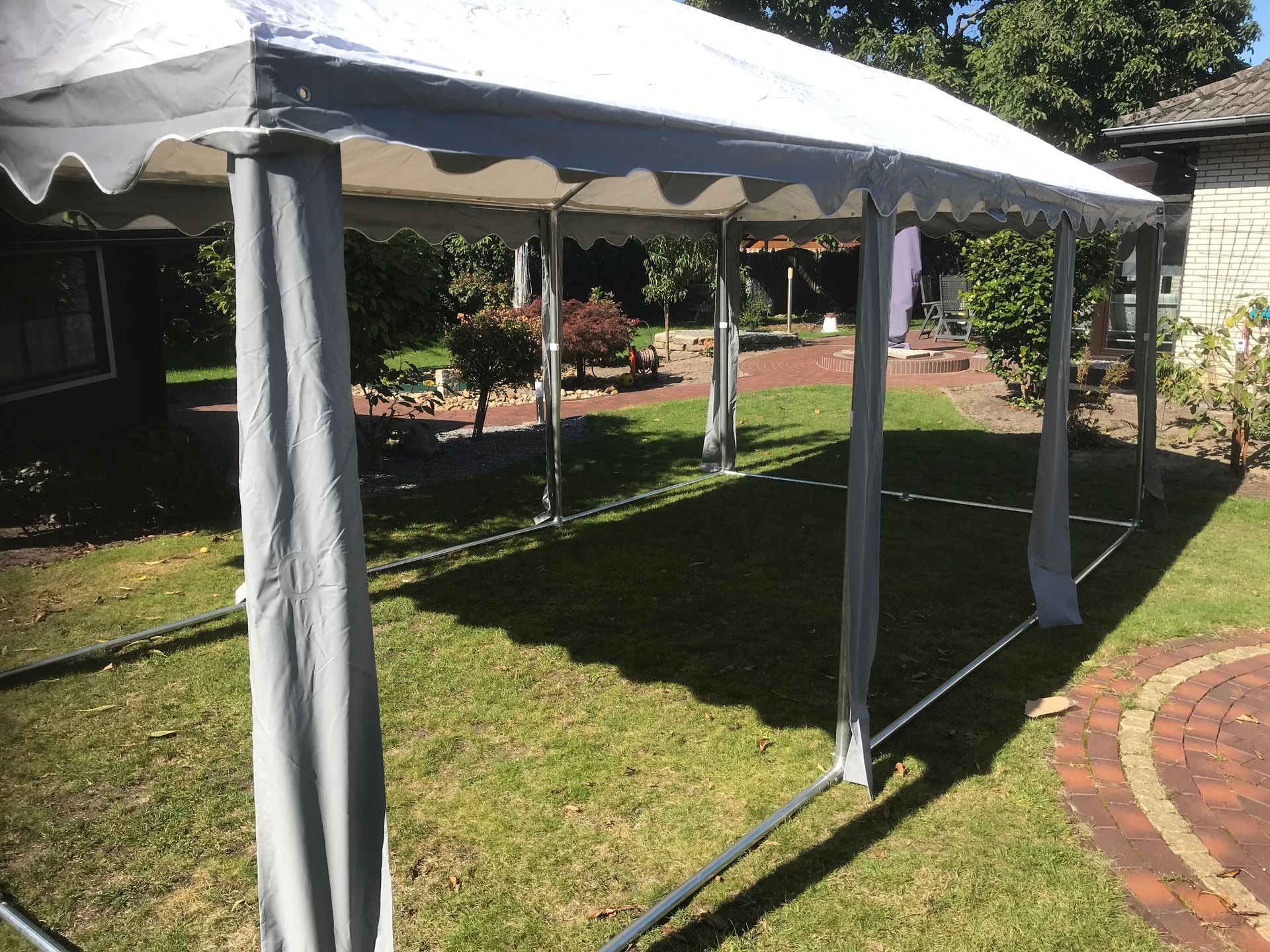 Auf einem grünen Rasen neben einem Backsteinweg steht ein weißes Festzelt mit grauen Eckstoffabdeckungen.