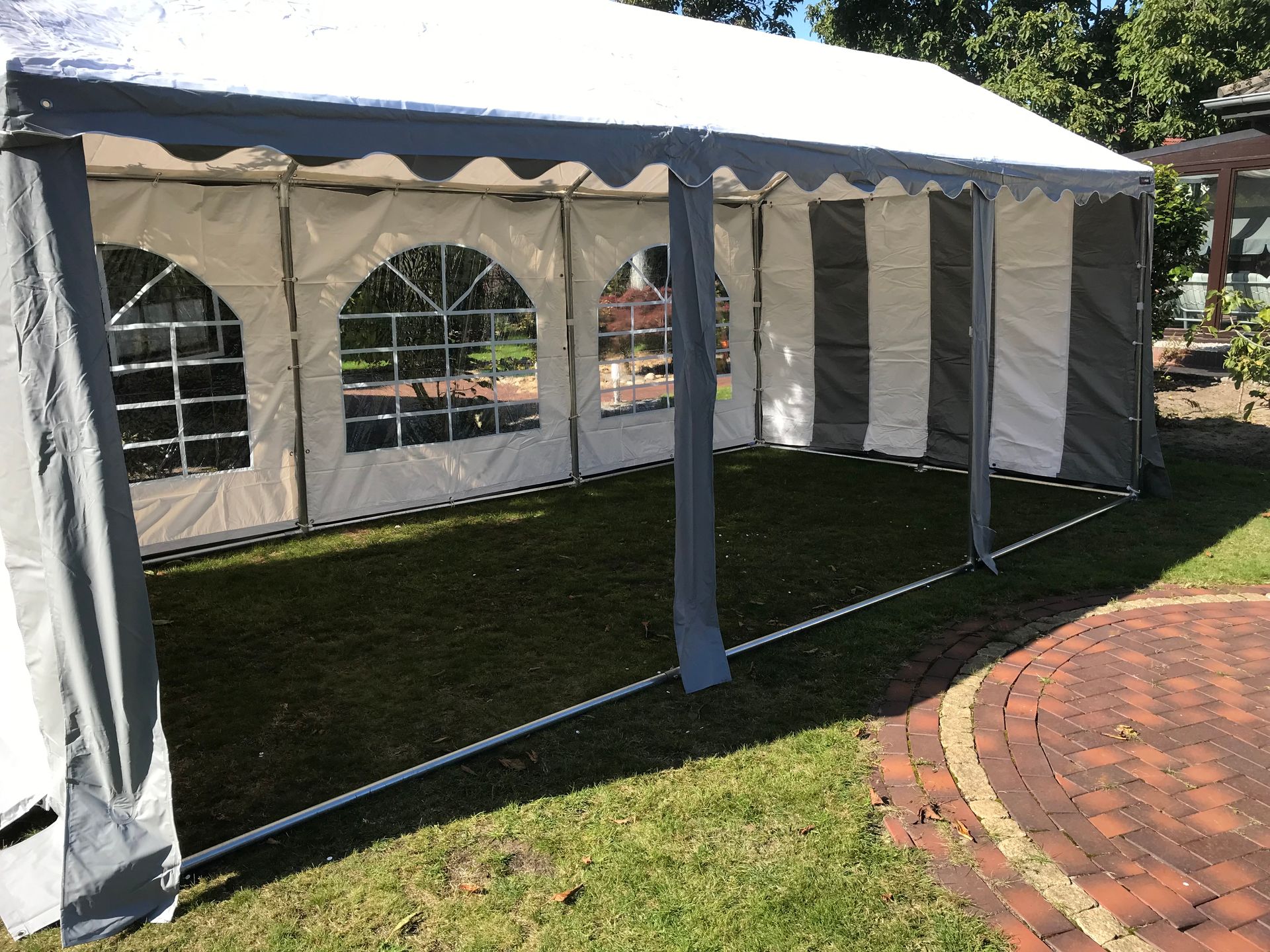 Ein weiß-graues Partyzelt mit verglasten Seitenwänden, aufgestellt auf einer Rasenfläche neben einer gemauerten Terrasse.