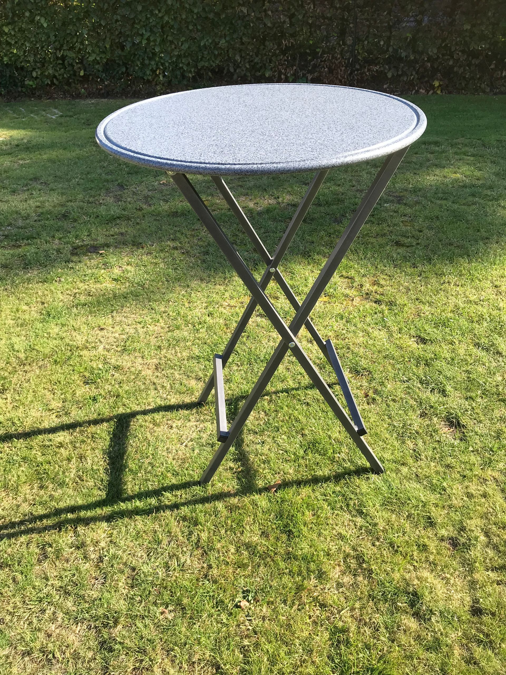 Auf einem grünen Rasen steht ein grau gesprenkelter runder Bistrotisch mit einem dunklen, klappbaren X-förmigen Metallgestell.