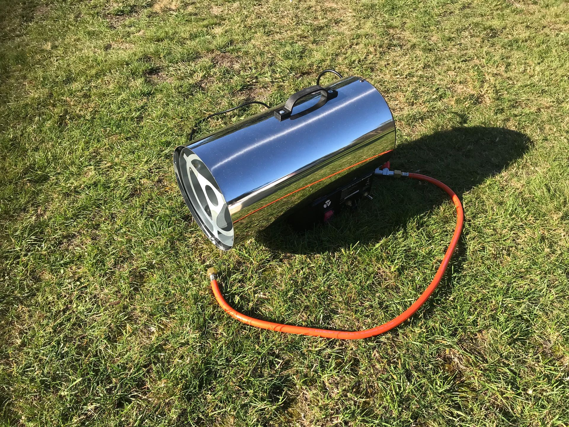 Auf einem grünen Rasen steht ein Propangas-Heißluftofen aus Edelstahl, der an einen orangefarbenen Brennstoffschlauch angeschlossen ist.