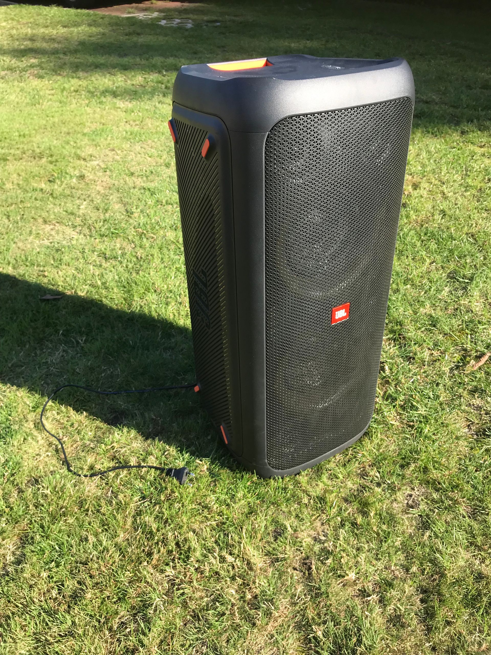 Ein schwarzer tragbarer JBL PartyBox-Lautsprecher mit orangefarbenen Akzenten steht im Sonnenlicht auf einer Rasenfläche.
