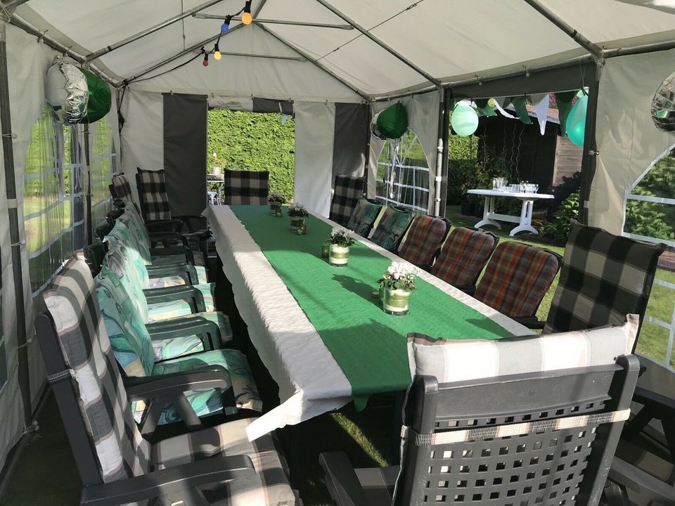 In einem weißen Baldachinzelt steht ein langer Tisch mit grüner Tischdecke und Blumengestecken, umgeben von Stühlen.